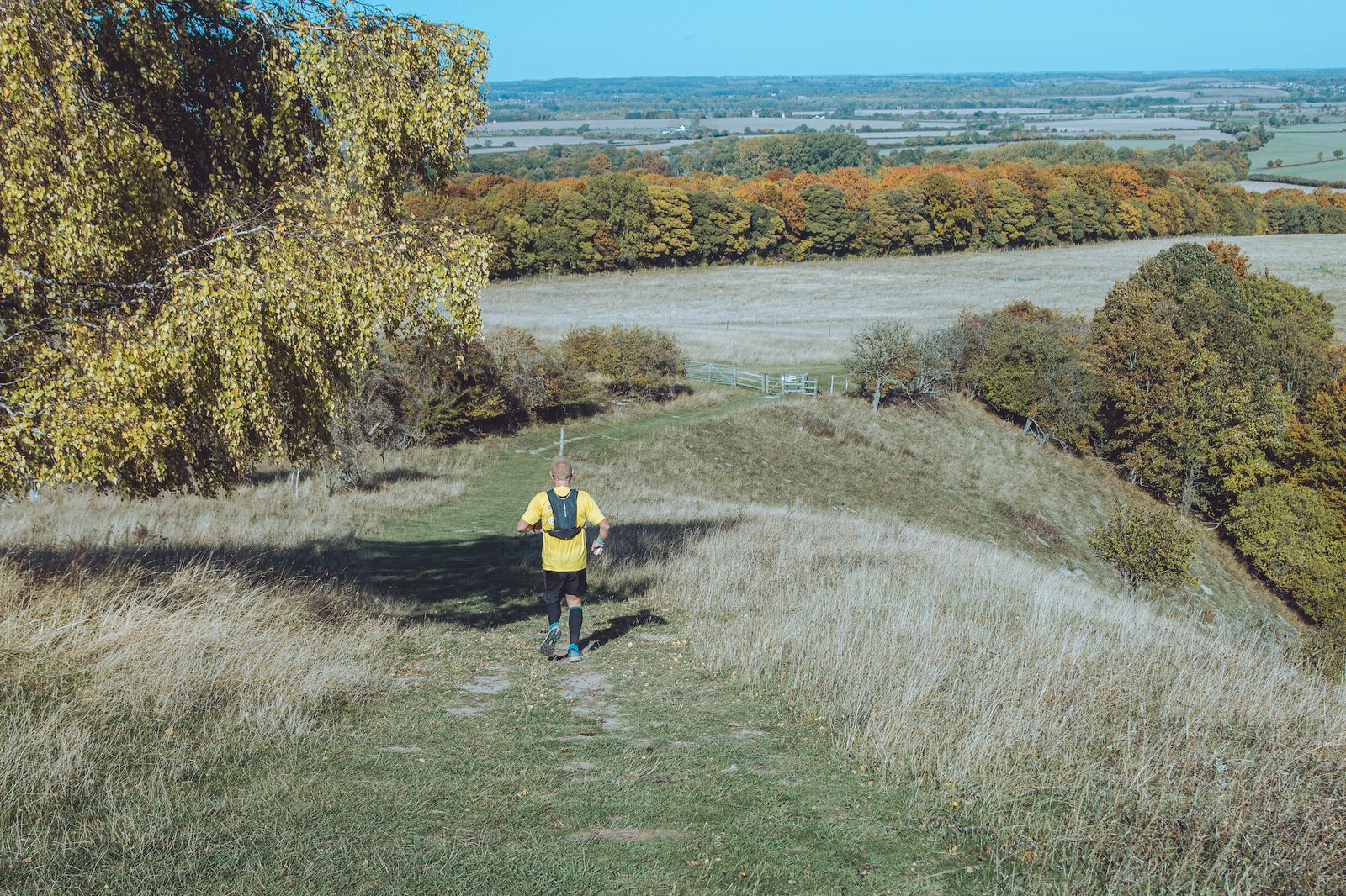 A person wearing a yellow vest jogs on a trail through a scenic landscape of rolling hills and autumnal trees. The sky is clear and the horizon stretches far in the distance, showcasing fields and patches of colorful foliage.