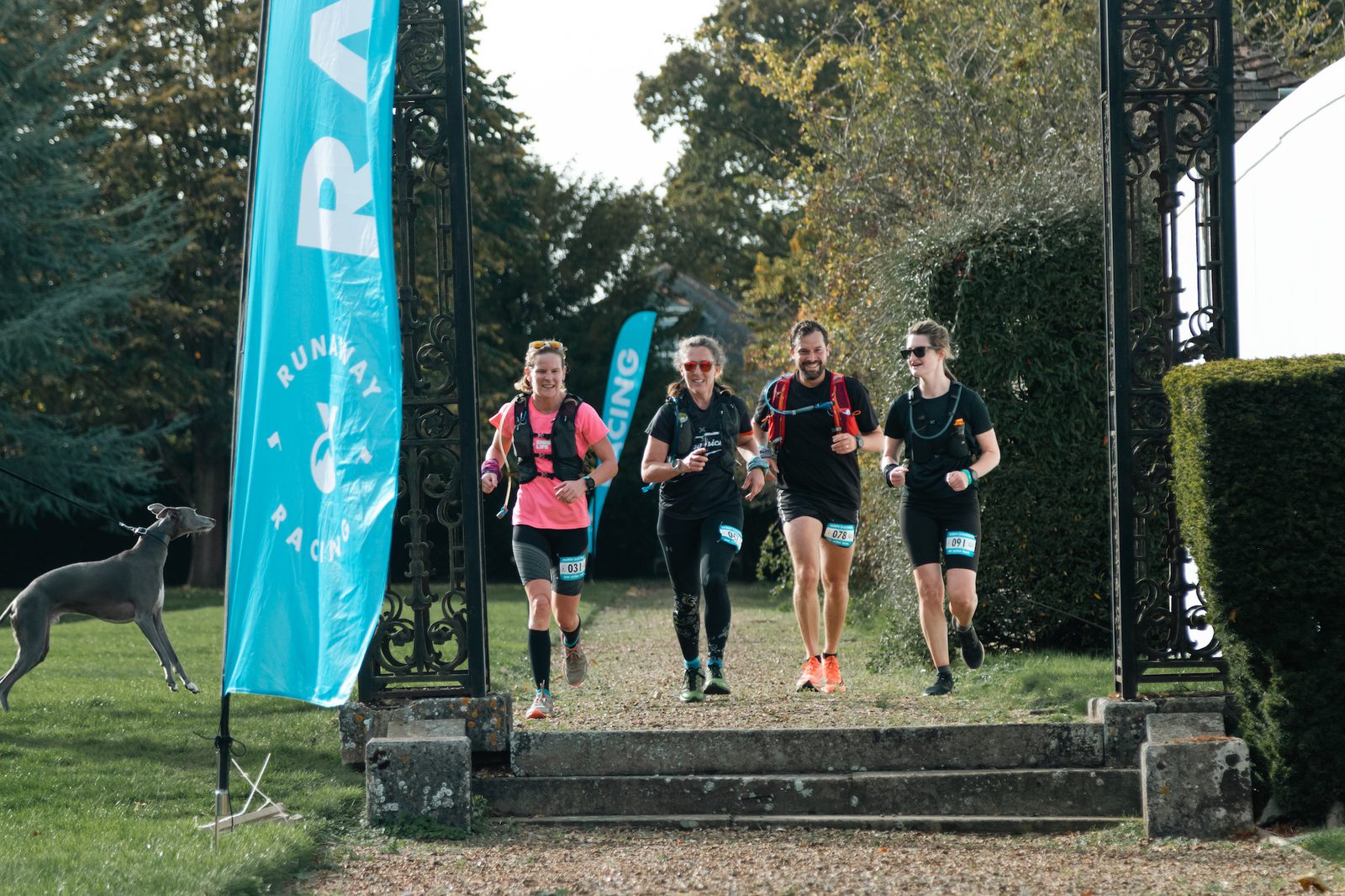 A group of four runners, three men and one woman, cross the finish line during a race. They are smiling and seem happy about their accomplishment. A banner with the word "RACING" is visible on the left side, and a dog is seen running alongside them.