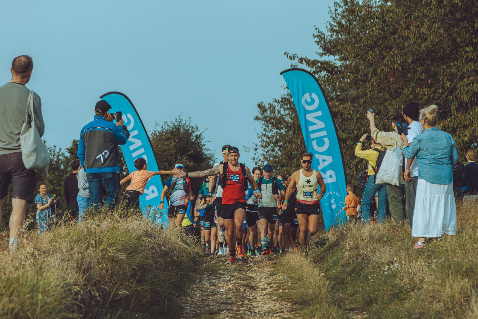 A group of runners, led by a man in a red tank top, competes on a dirt trail flanked by tall grass and trees. Spectators cheer them on, standing near blue banners with white text. The sky is clear and sunny, adding to the lively atmosphere of the race event.