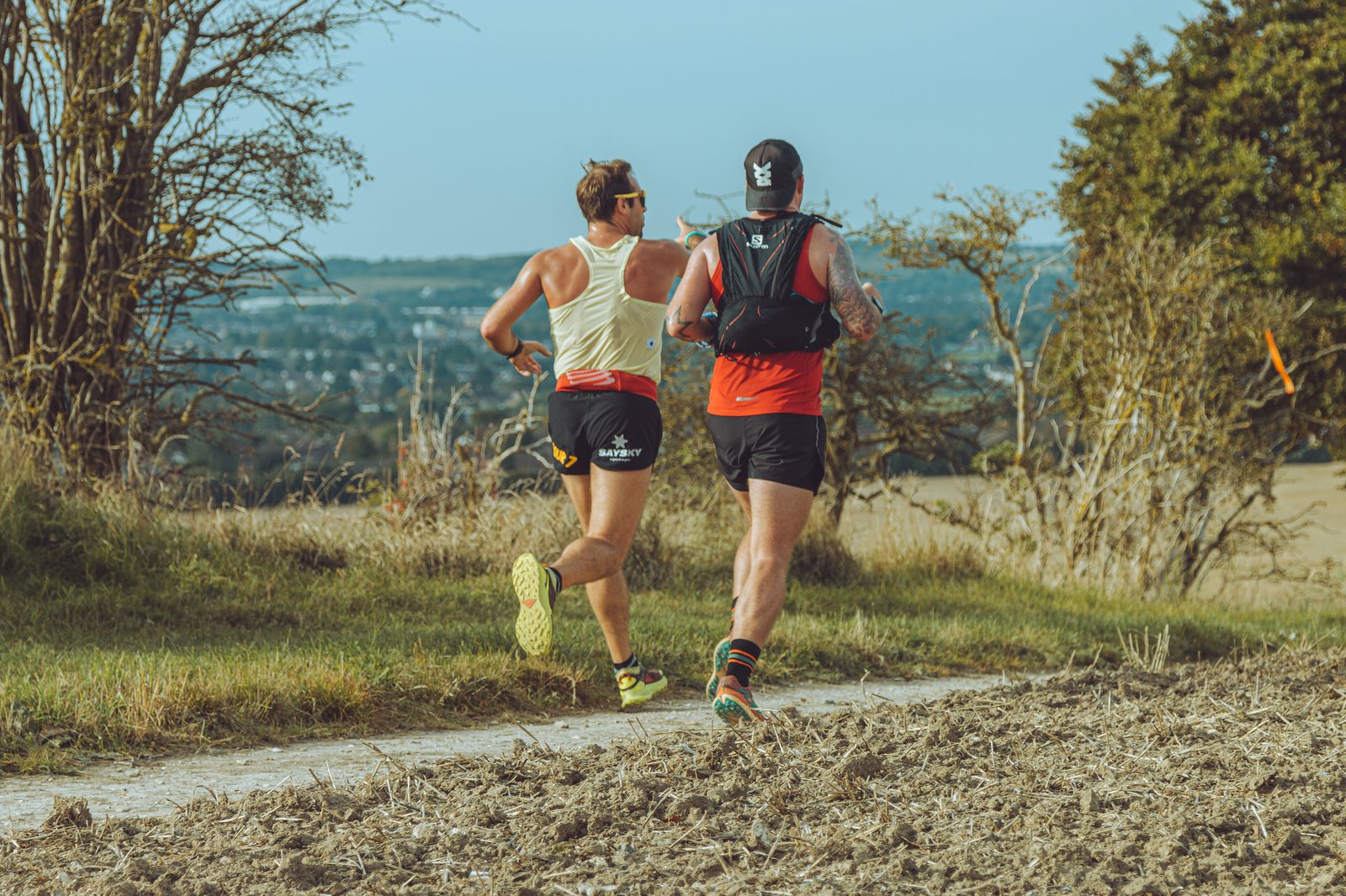Two runners are photographed from behind as they run along a dirt path through a rural landscape. One wears a white tank top and the other a red one; both have black shorts. Trees and fields surround them, with a town in the background under a clear blue sky.