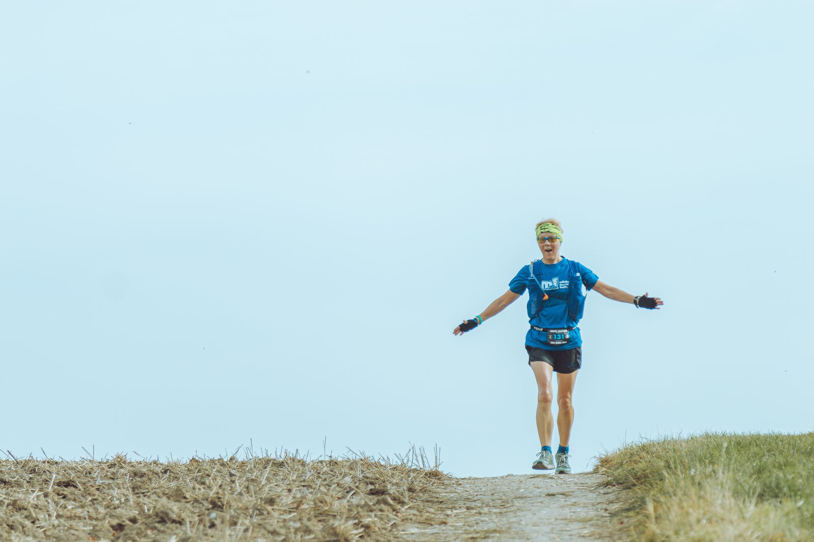 A person running along an outdoor trail on a clear day. They are wearing a blue shirt, black shorts, a green headband, and blue running shoes. Their arms are extended outward, and a fanny pack is strapped around their waist. The background is an open, grassy area.