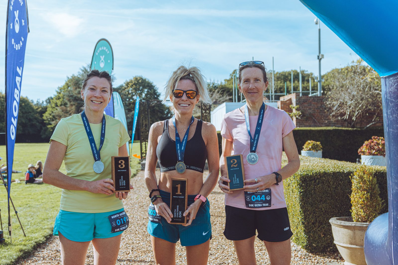 Three women stand under a blue archway outdoors, holding awards and wearing medals around their necks. The woman on the left holds a plaque with "3," the woman in the middle holds a plaque with "1," and the woman on the right holds a plaque with "2.