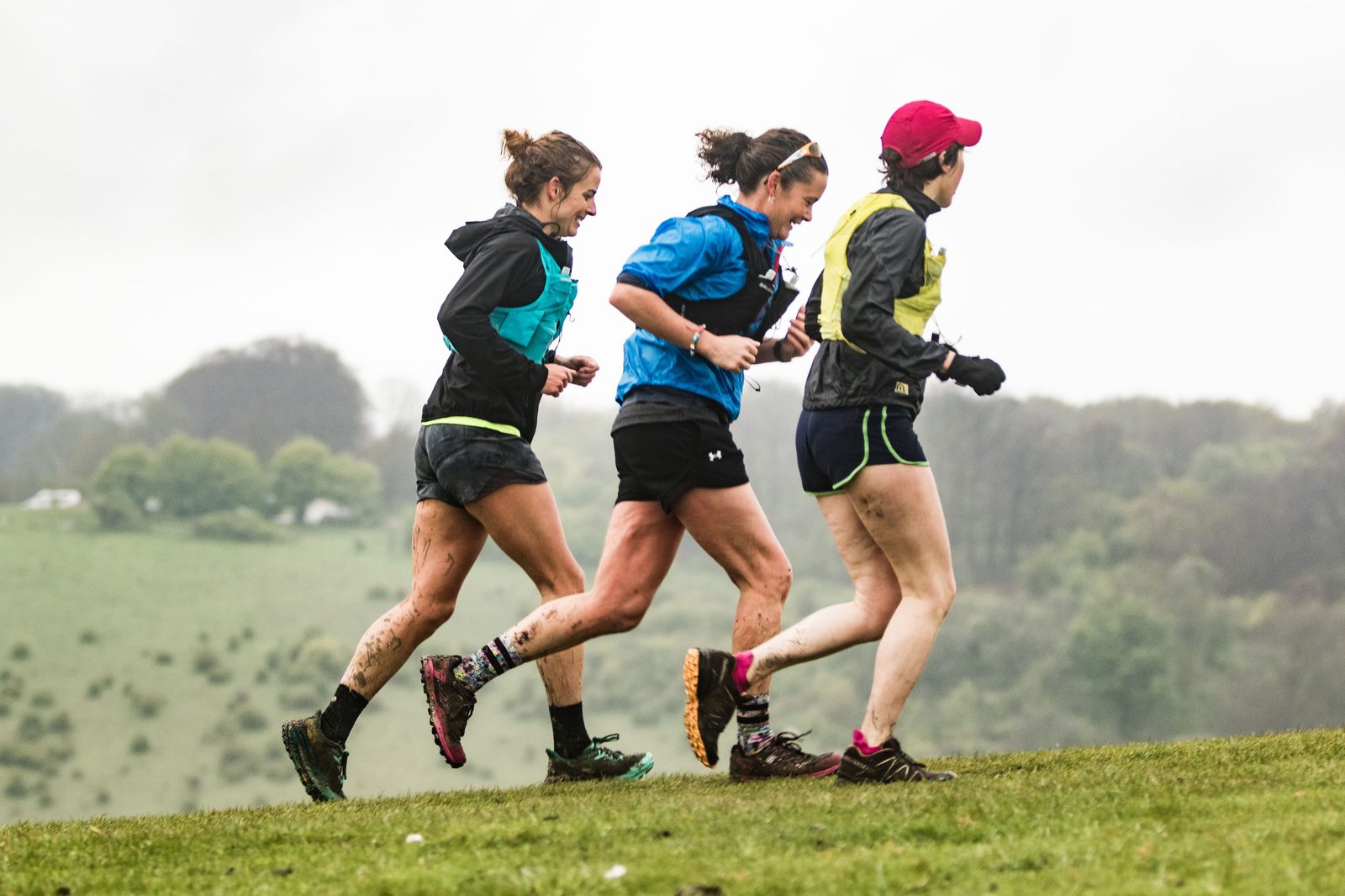 Three people are running on a grassy hill in athletic gear. They appear muddy, suggesting they are participating in an off-road or trail race. The sky is overcast, and there are trees in the background. One person wears a pink cap, another a blue jacket, and the third a black hoodie.