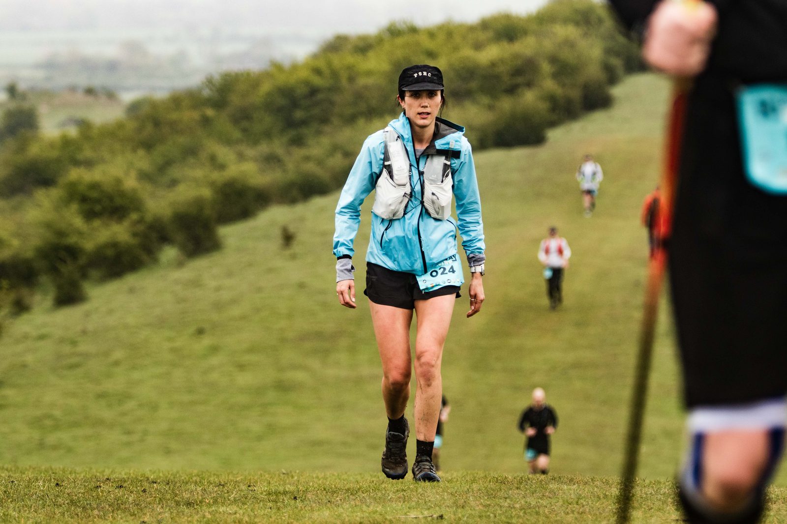 A person wearing a blue jacket, white running vest, black shorts, and a black cap is participating in an outdoor race on a grassy trail. They are approaching the camera, with more runners visible in the background on a slight incline.