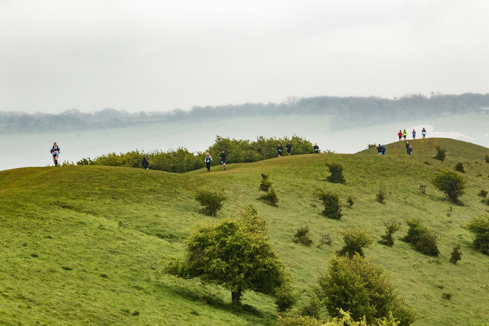 A misty landscape with rolling green hills and scattered small trees. Several people are walking along the hilltops, spaced apart, wearing jackets and carrying backpacks. In the distance, there is a body of water partially obscured by fog.