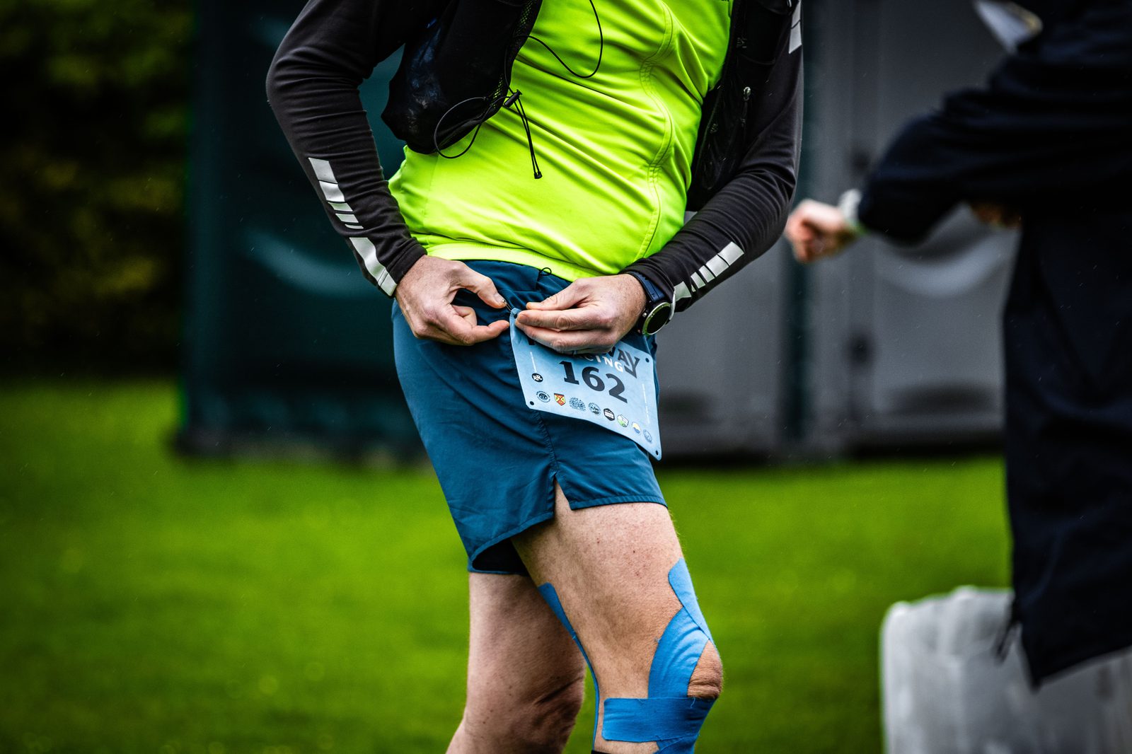 A person wearing a bright yellow-green shirt and blue shorts with a race bib numbered 162 adjusts their clothing. The person also has blue kinesiology tape on their knee. Another person partially enters the frame on the right side. They are outside on grass.