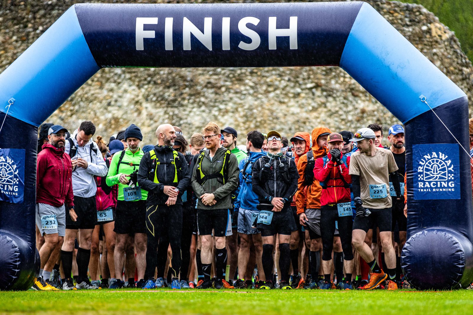 A group of runners stands at the starting line beneath a large inflatable arch with the word "FINISH" written on it. The runners are wearing various colorful athletic gear, and the atmosphere appears tense and focused as they prepare for the race.