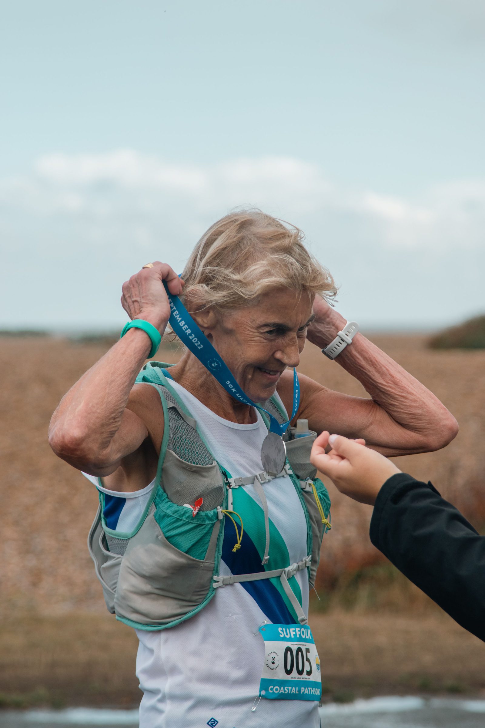 An elderly woman with short gray hair is putting a blue medal around her neck. She is wearing a light gray hydration vest over a white and blue tank top, and a blue race bib with the number "005". Another person extends a hand towards her. The background shows a rocky and grassy terrain.