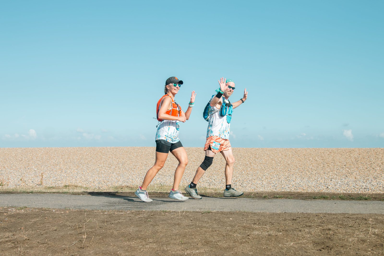 Two people in athletic gear are walking briskly on a paved path beside a rocky shore under a clear blue sky. Both are wearing sunglasses, caps, and smartwatches. They wave and smile toward the camera, appearing cheerful and energetic.