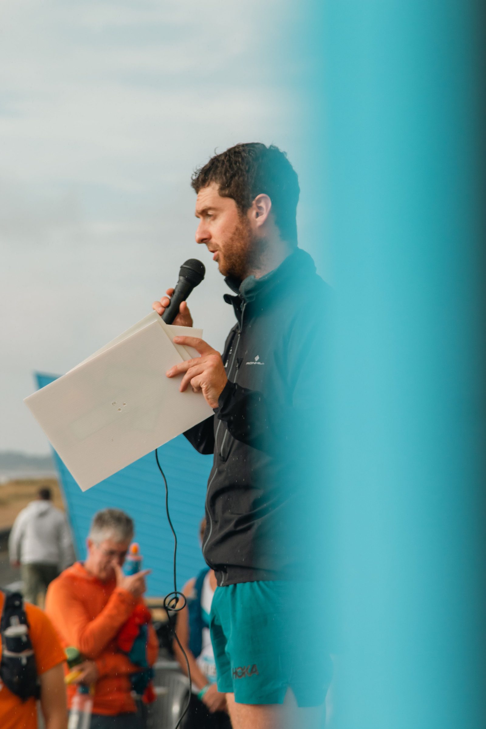 A bearded man in a black jacket and green shorts holds a microphone and a piece of paper while speaking to a crowd. The background features a blue structure and another person in an orange shirt. A blue blur partially obstructs the view.