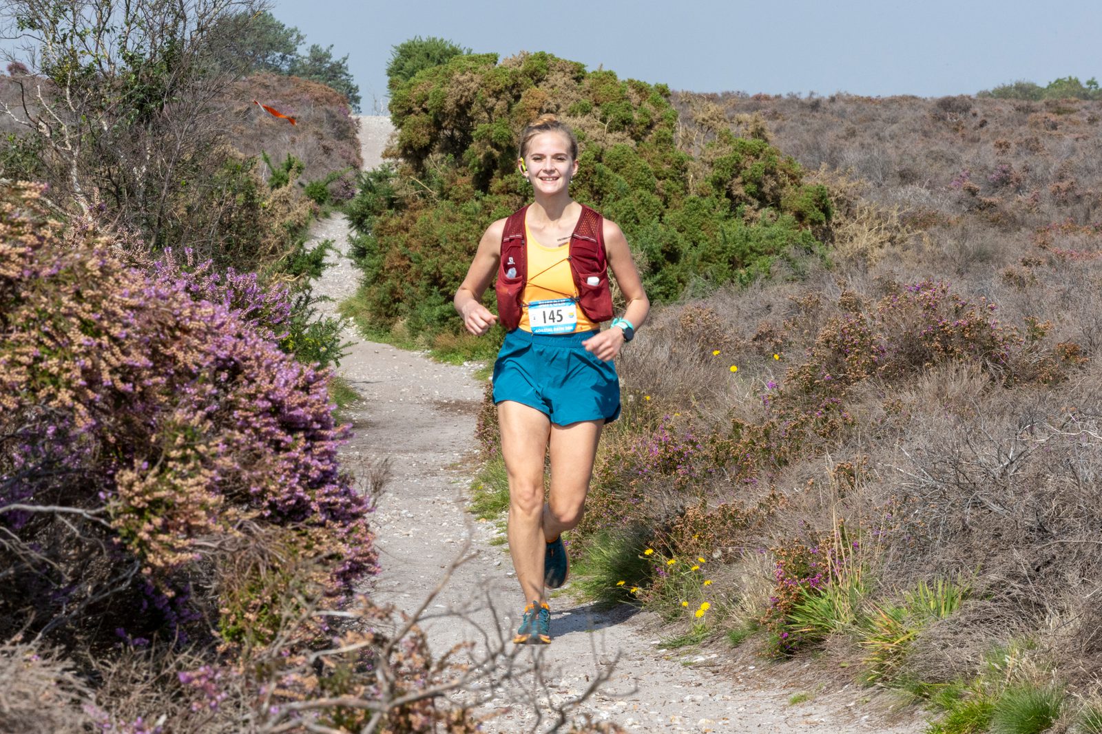 A person with a race bib number 145, wearing a maroon top, blue shorts, and a maroon hydration pack, is running on a trail surrounded by bushes and wildflowers. The sky is clear and blue. The runner appears to be smiling and enjoying the run.