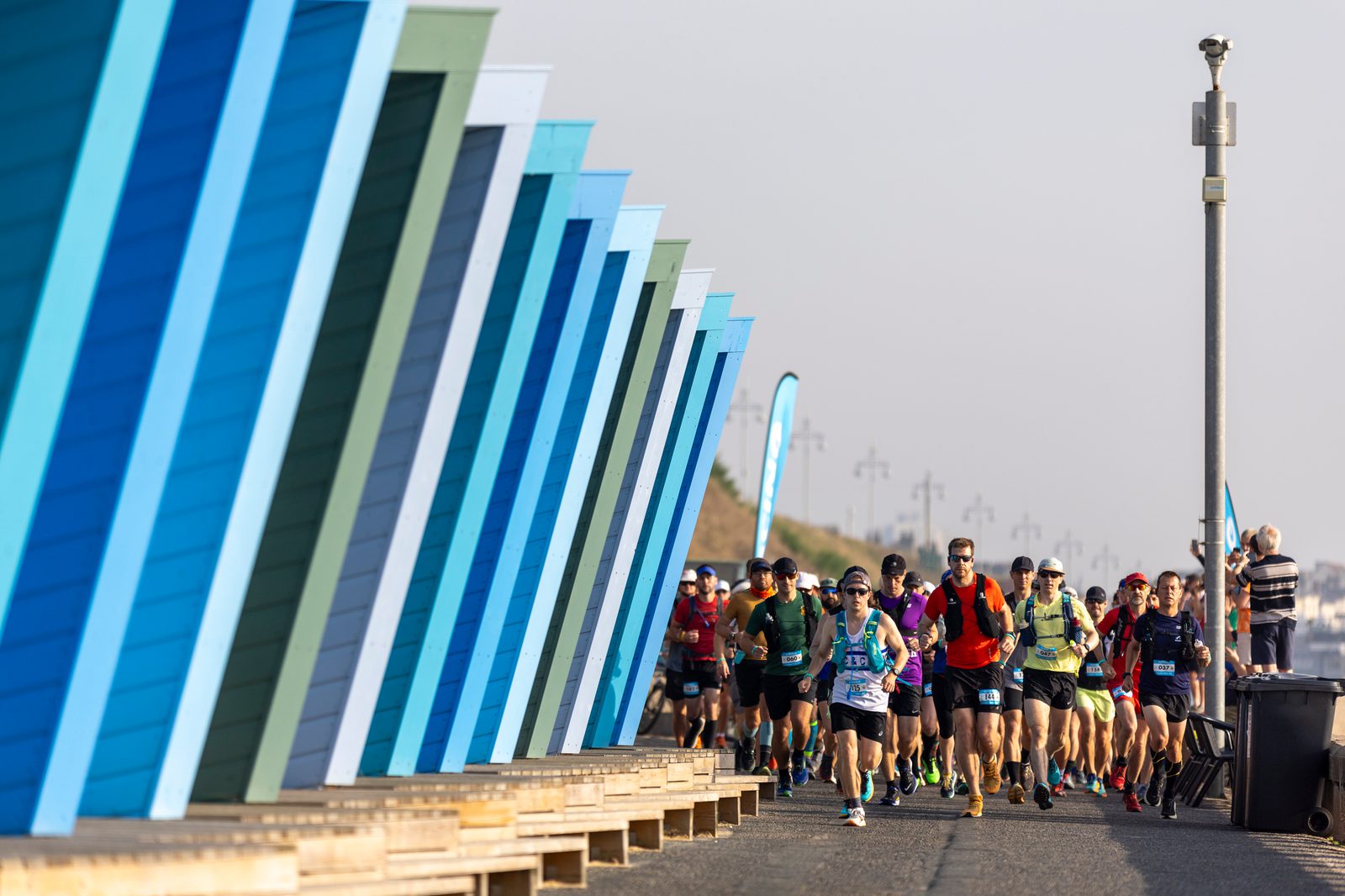 Runners participating in a marathon race along a scenic beachside route, with colorful angular beach huts lining one side of the path. The runners, dressed in athletic gear, progress under a clear blue sky. The backdrop shows lampposts and the vast ocean.