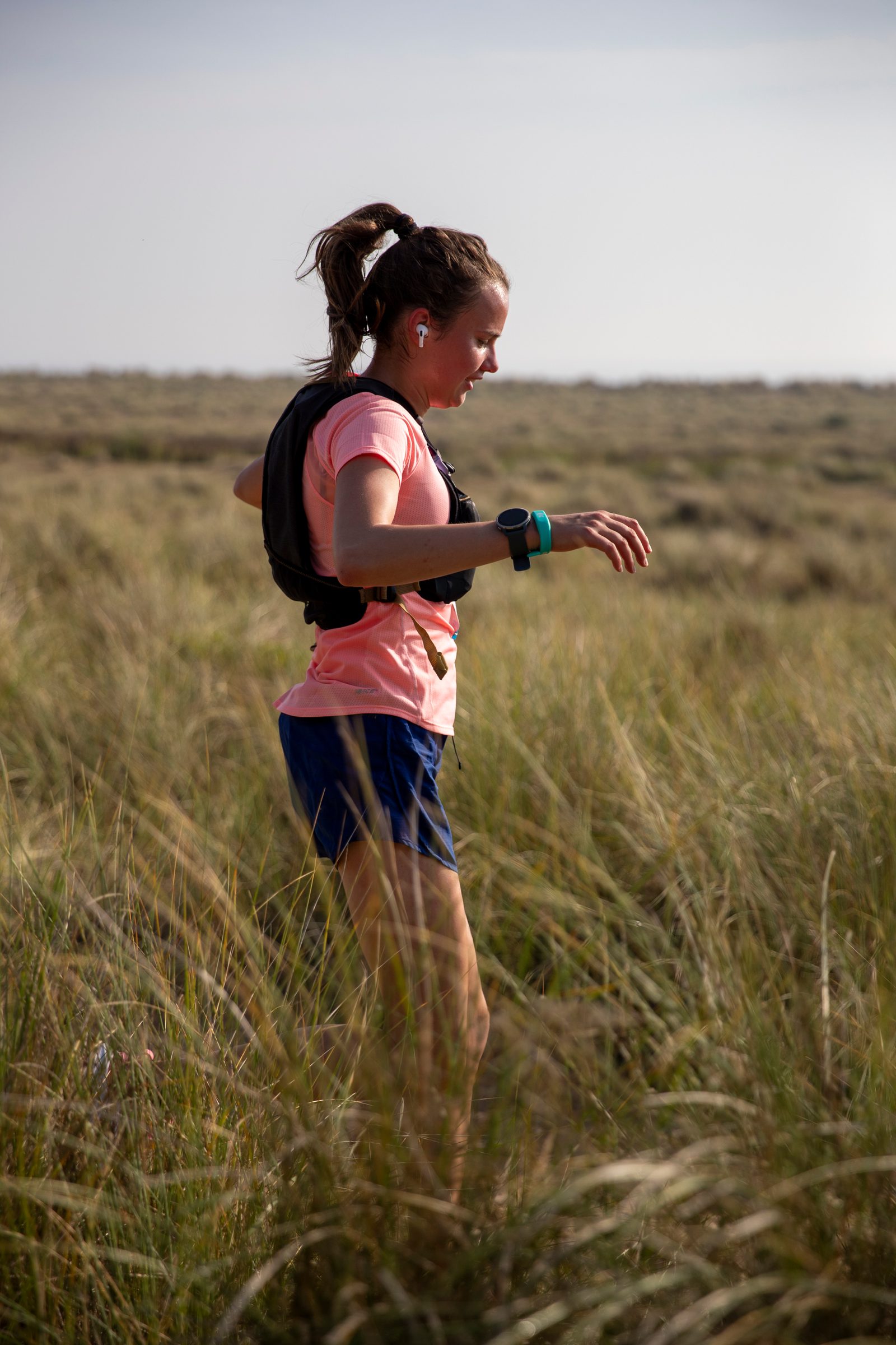 A person with a ponytail, wearing a pink shirt, blue shorts, a black hydration vest, and a wristband, is running through tall grass in an open field. The person is facing to the right and appears to be in motion. The sky is clear.