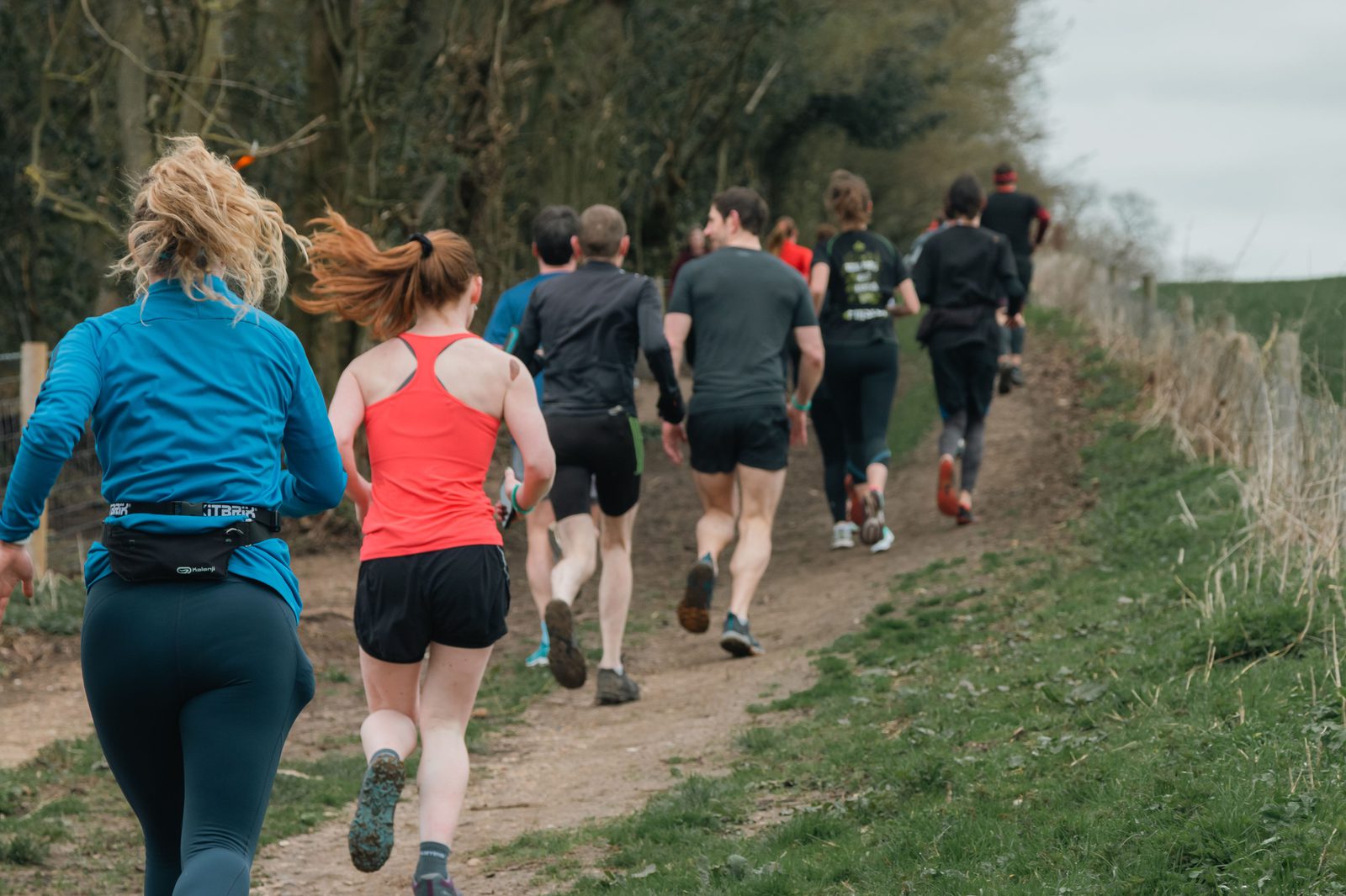 A group of people running along a forest trail. Most of the runners are dressed in athletic wear, and some appear to be in mid-stride. The path runs uphill and is surrounded by trees and grass. The sky is overcast, suggesting a cool day.