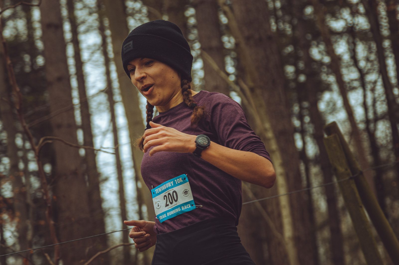 A woman, wearing a black beanie and a purple long-sleeve shirt, runs through a forest during a race. She wears a race bib numbered 200 and has braided hair. She appears focused and determined as she navigates the wooded trail.