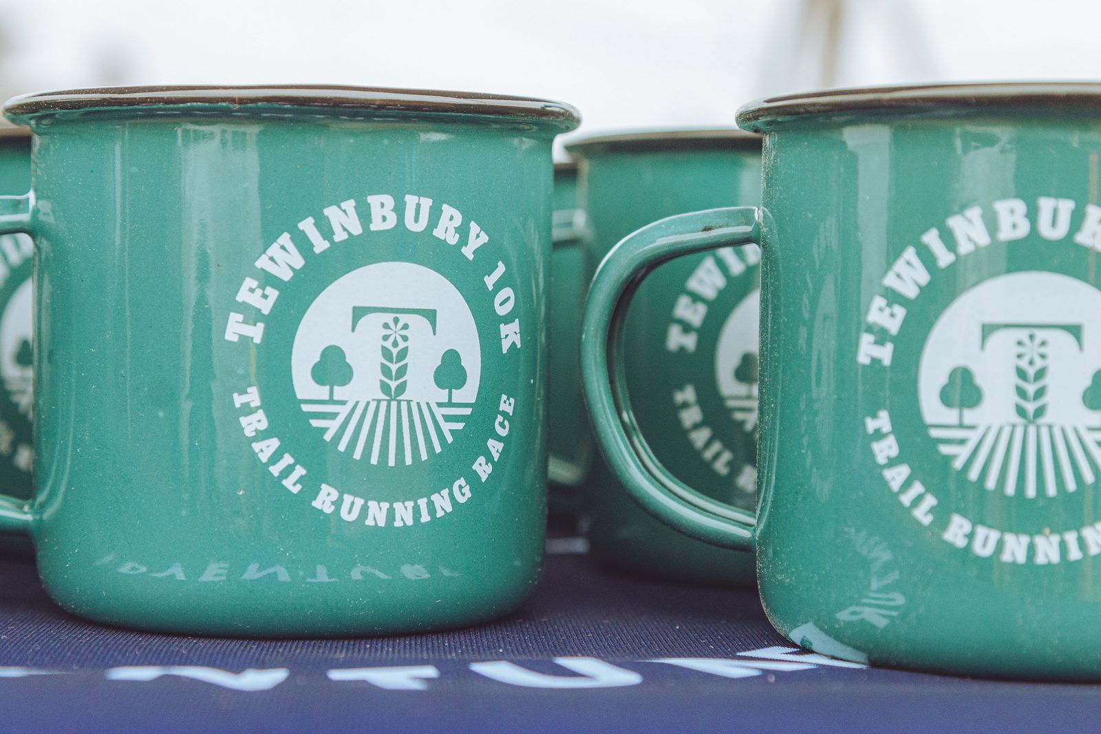 A close-up photo of green metal mugs featuring a white logo and text that reads, "TEWINBURY 10K TRAIL RUNNING RACE." The mugs are placed on a dark surface, suggesting they may be souvenirs or prizes for participants of the event.