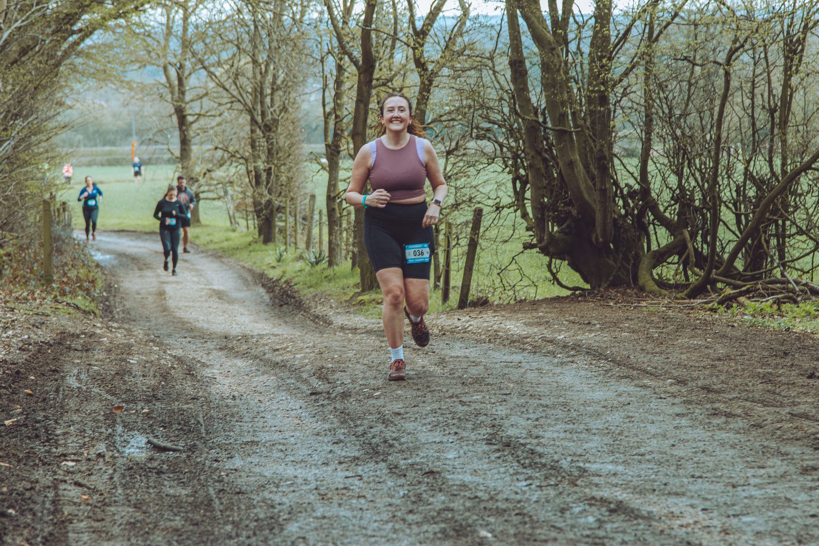 A person wearing a purple tank top and black shorts is running down a dirt path through a wooded area, smiling. Several other runners can be seen in the background. The sky is overcast, and the scene has a serene, natural setting.