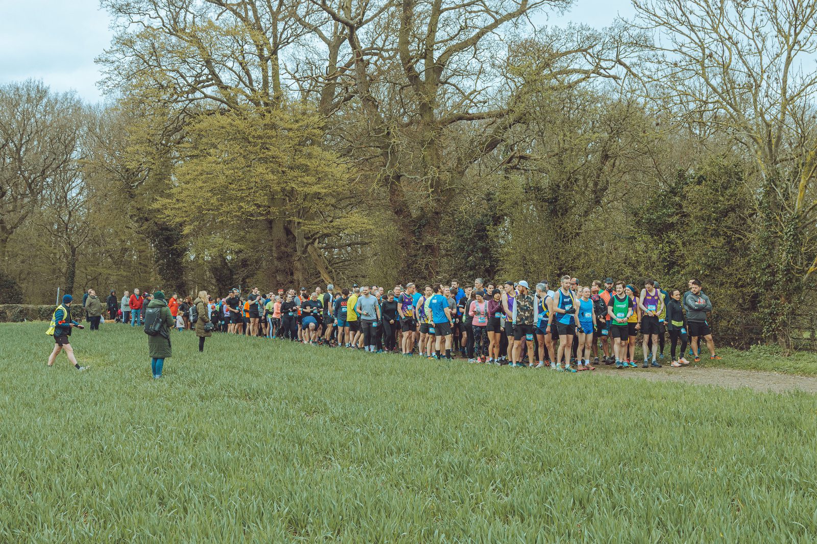 A large group of runners in athletic gear stand at the starting line of a race on a grassy field surrounded by trees. Two people in reflective clothing stand nearby, likely organizing the event, while a third person walks away from the group.