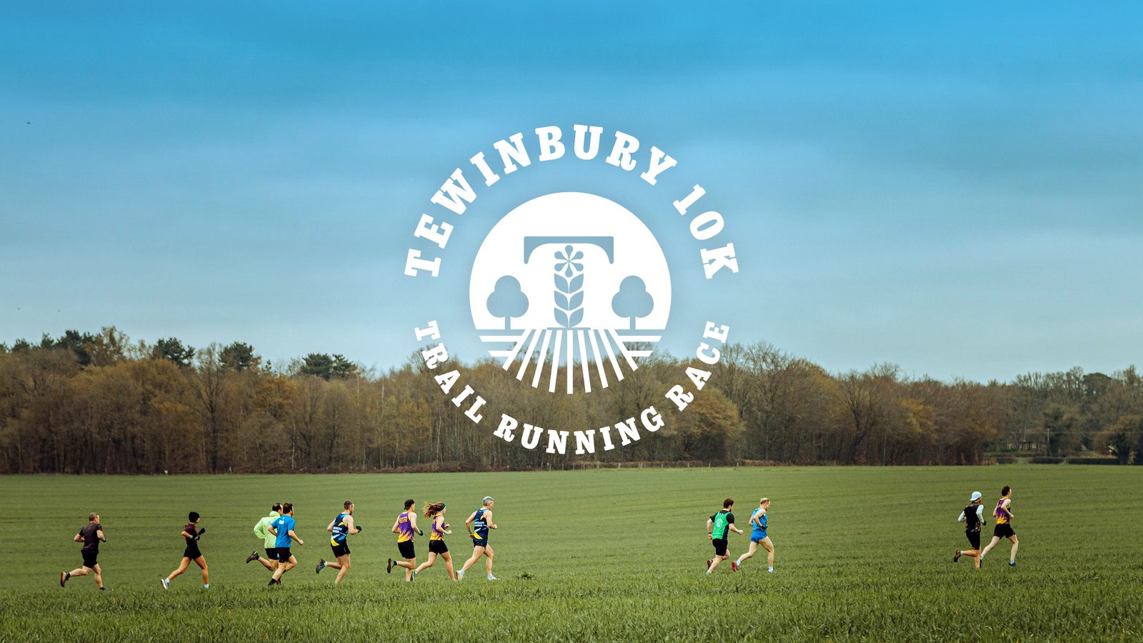 A group of runners participating in the Tewinbury 10K Trail Running Race runs across a green field. The race logo overlays the image, depicting a tree and two smaller trees under the event name. The sky is clear and blue, contrasting with the greenery below.