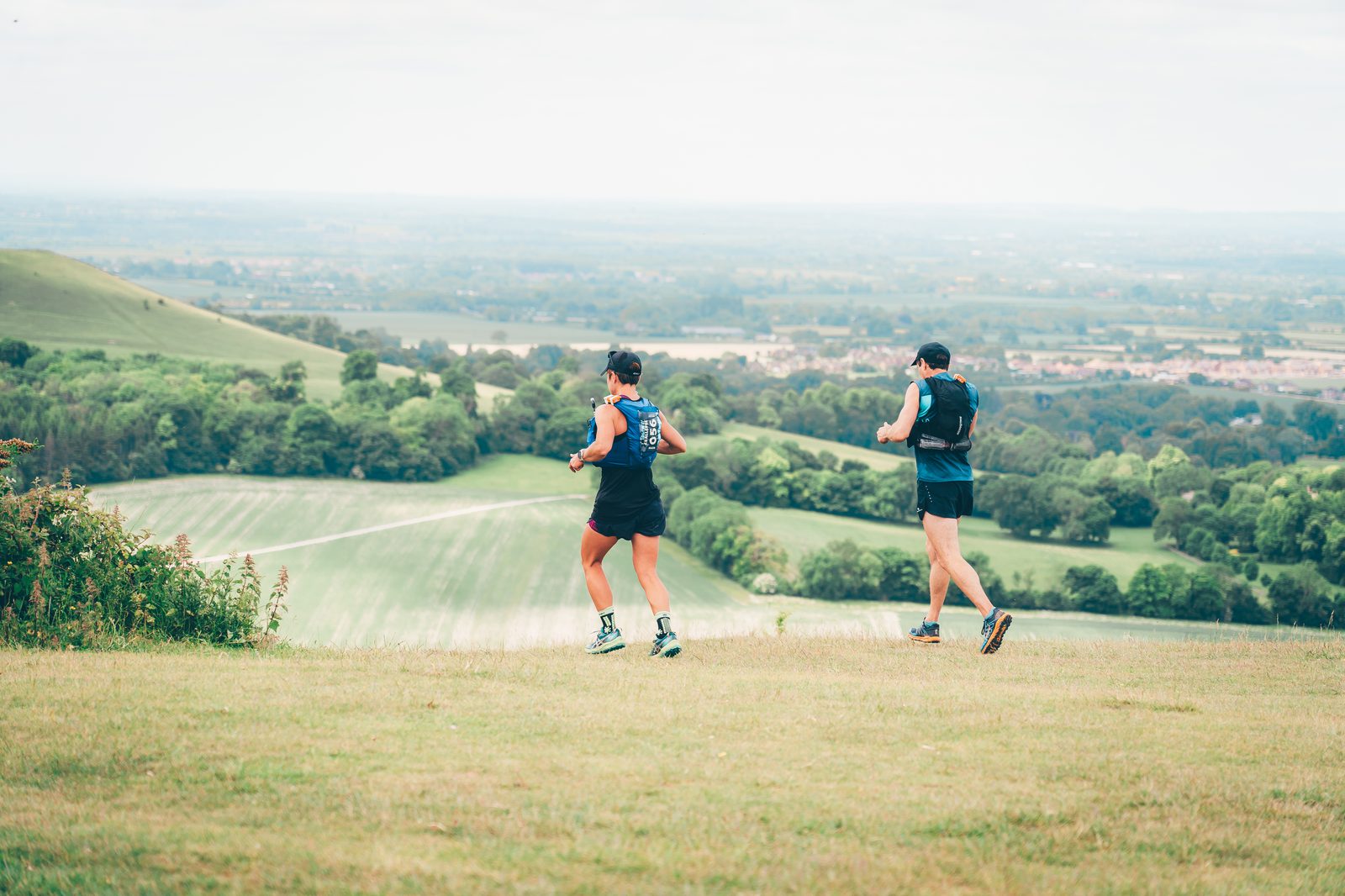 Two people in athletic gear run across a grassy hilltop with lush green fields, trees, and distant villages visible in the background under a cloudy sky.