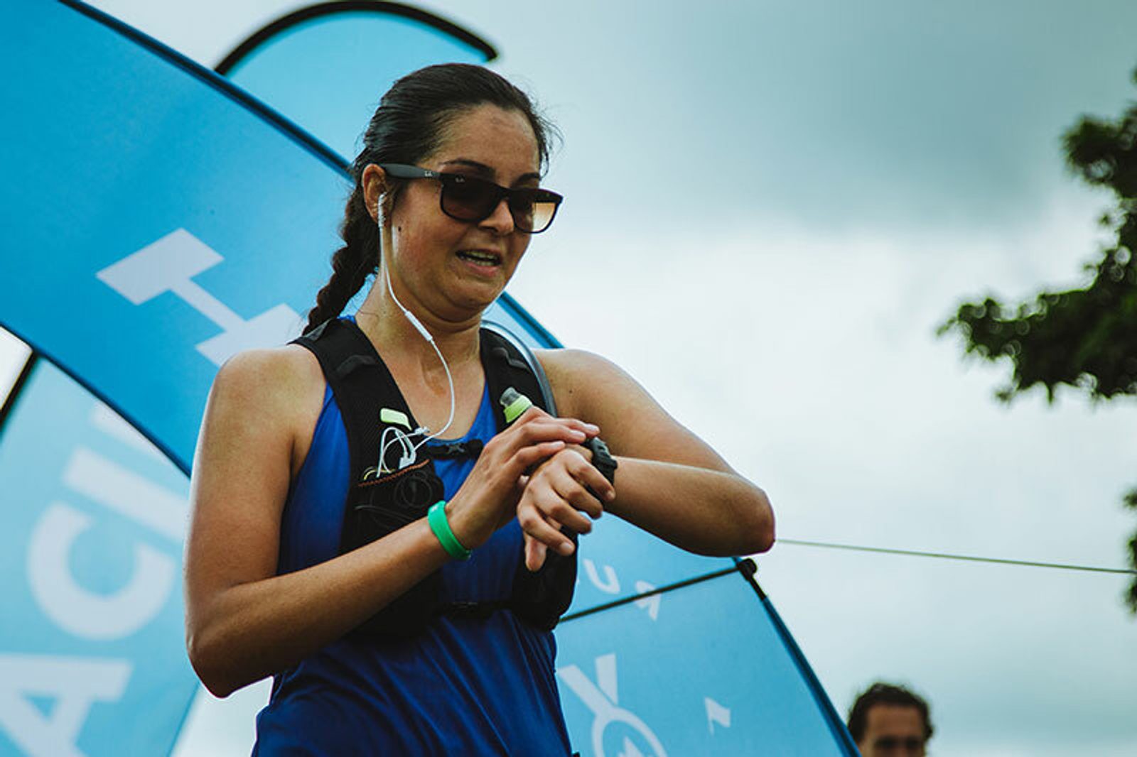 A woman wearing a blue tank top, sunglasses, and earphones looks at her watch while participating in an outdoor event. She has a hydration pack on her back, and there are blue event banners and a cloudy sky in the background.