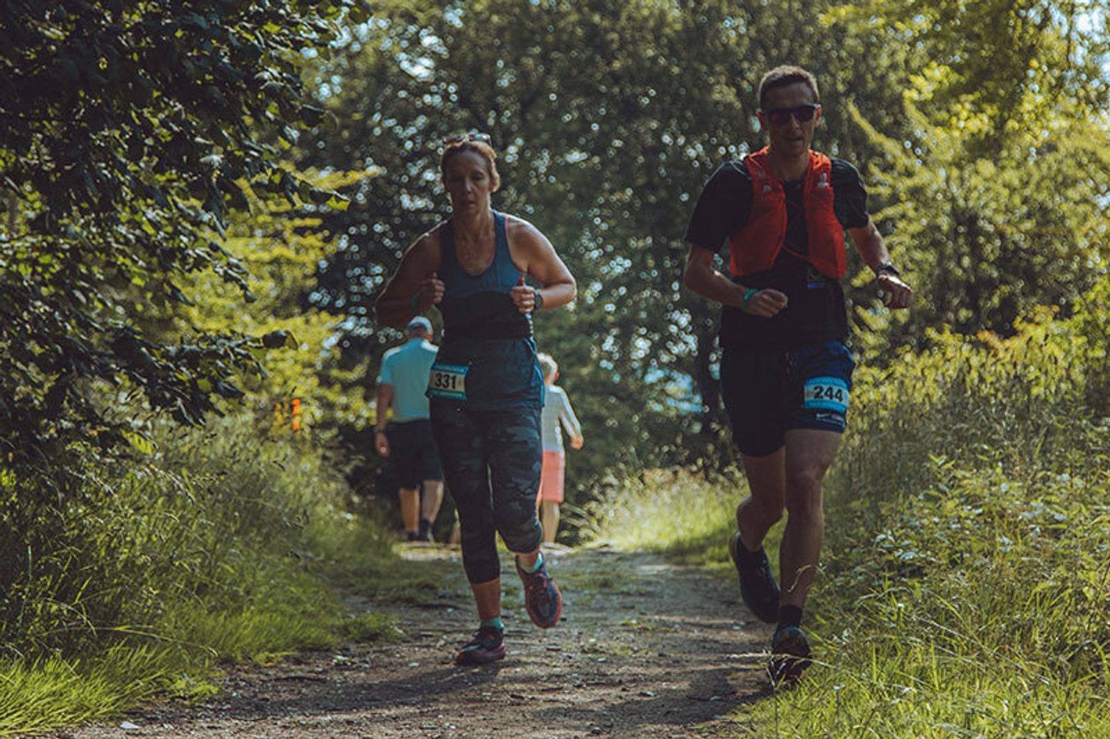 Two people are running on a forest trail with race bibs pinned to their clothing. The person on the left wears a tank top and leggings, and the person on the right wears a shirt with a red vest. Sunlight filters through the trees, illuminating the greenery.