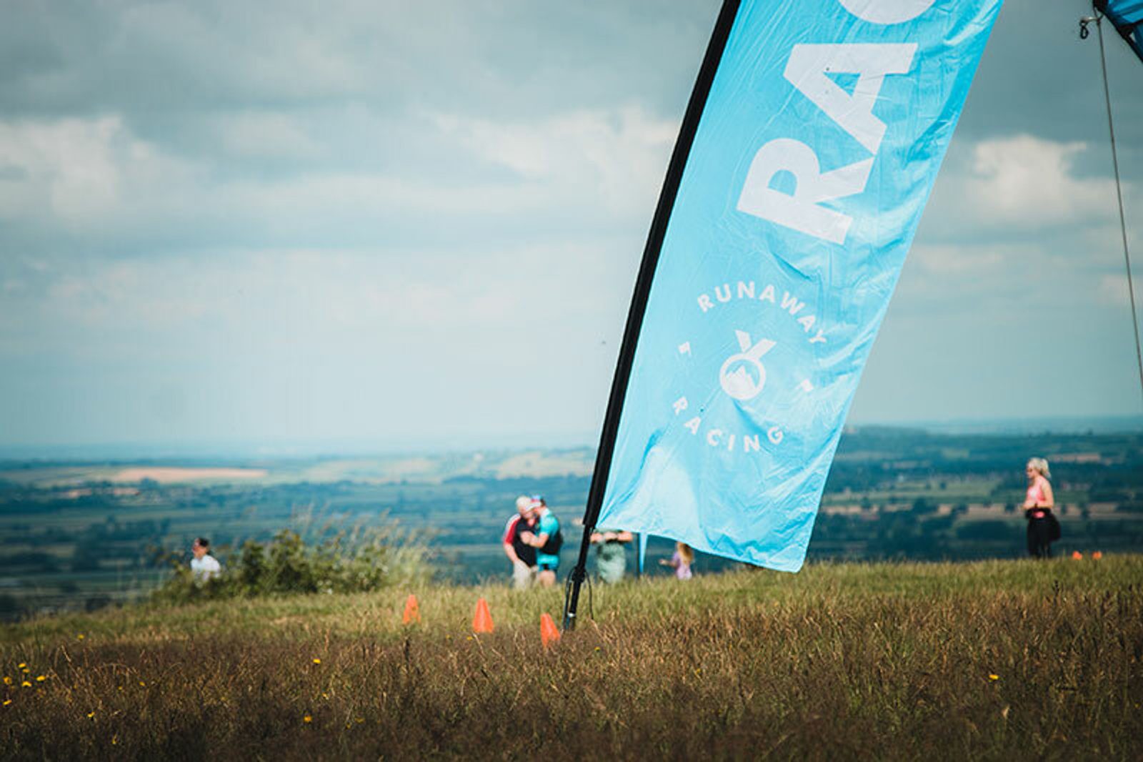 A blue flag with the text "Runaway Racing" flutters on a grassy hill. Several people stand in the background near small orange cones, with a vast landscape of fields and a cloudy sky stretching out in the distance.