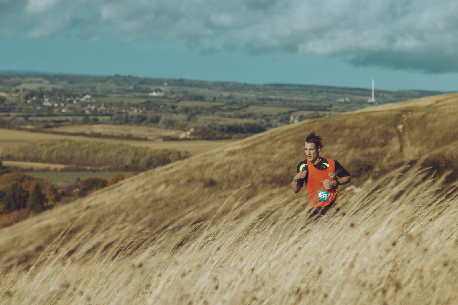 A person in an orange shirt and a race bib runs through a field of tall, dry grass on a hill. The landscape behind features rolling green fields, distant buildings, and a cloudy sky. The individual appears focused and determined.