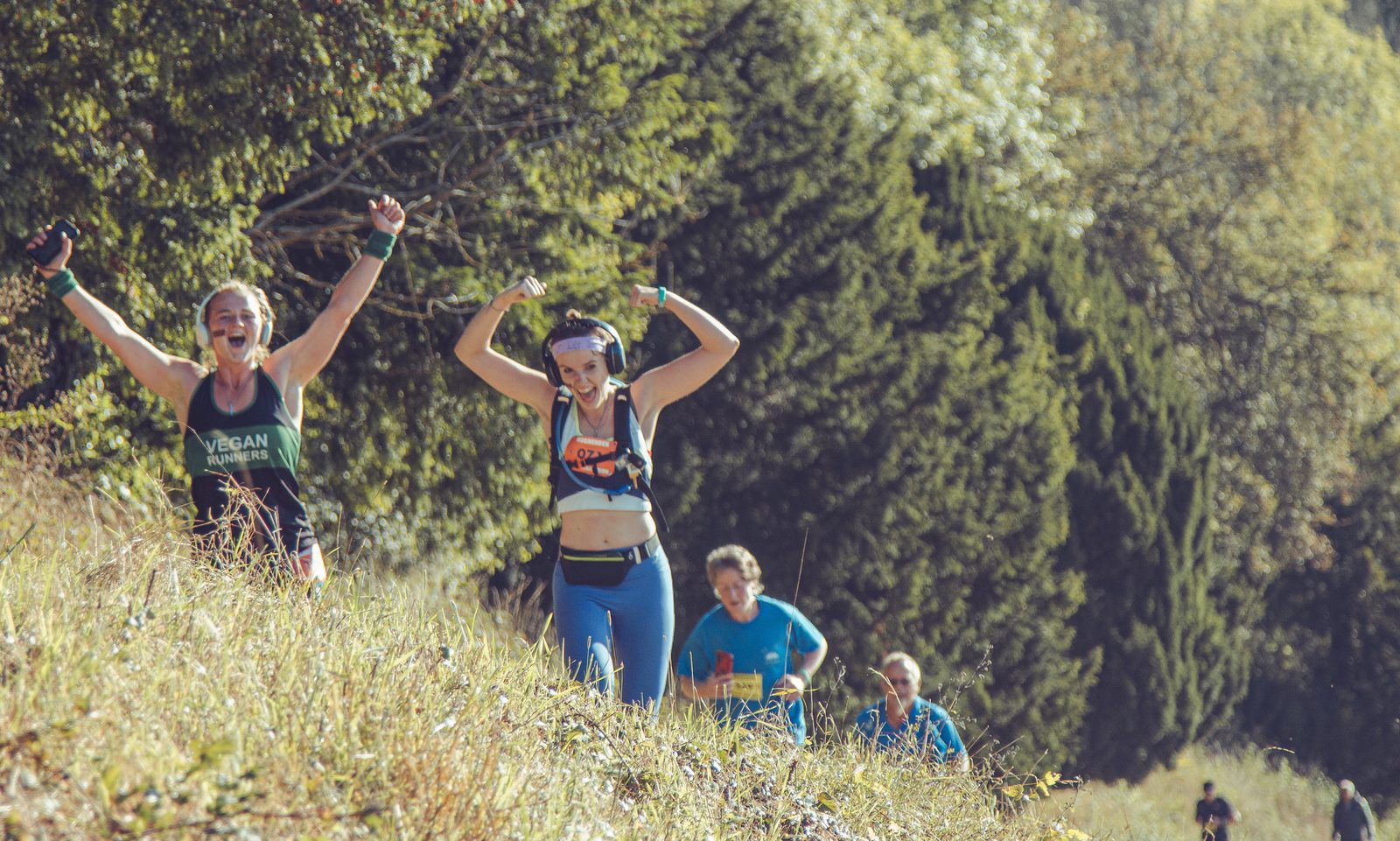 Four runners, two women in front and two men behind, are on a grassy, uphill trail surrounded by trees. The women smile and raise their arms triumphantly. The scene is sunny and energetic, suggesting a trail race or marathon.