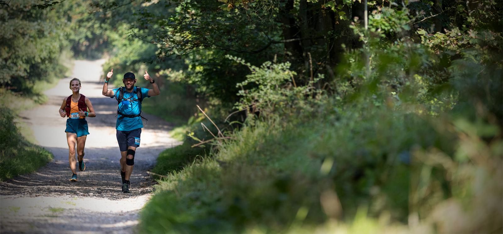Two people in athletic gear are running on a sunlit forest trail. The person in front wears a blue outfit and raises their arms in celebration, while the person behind in an orange shirt smiles, both enjoying the natural scenery.