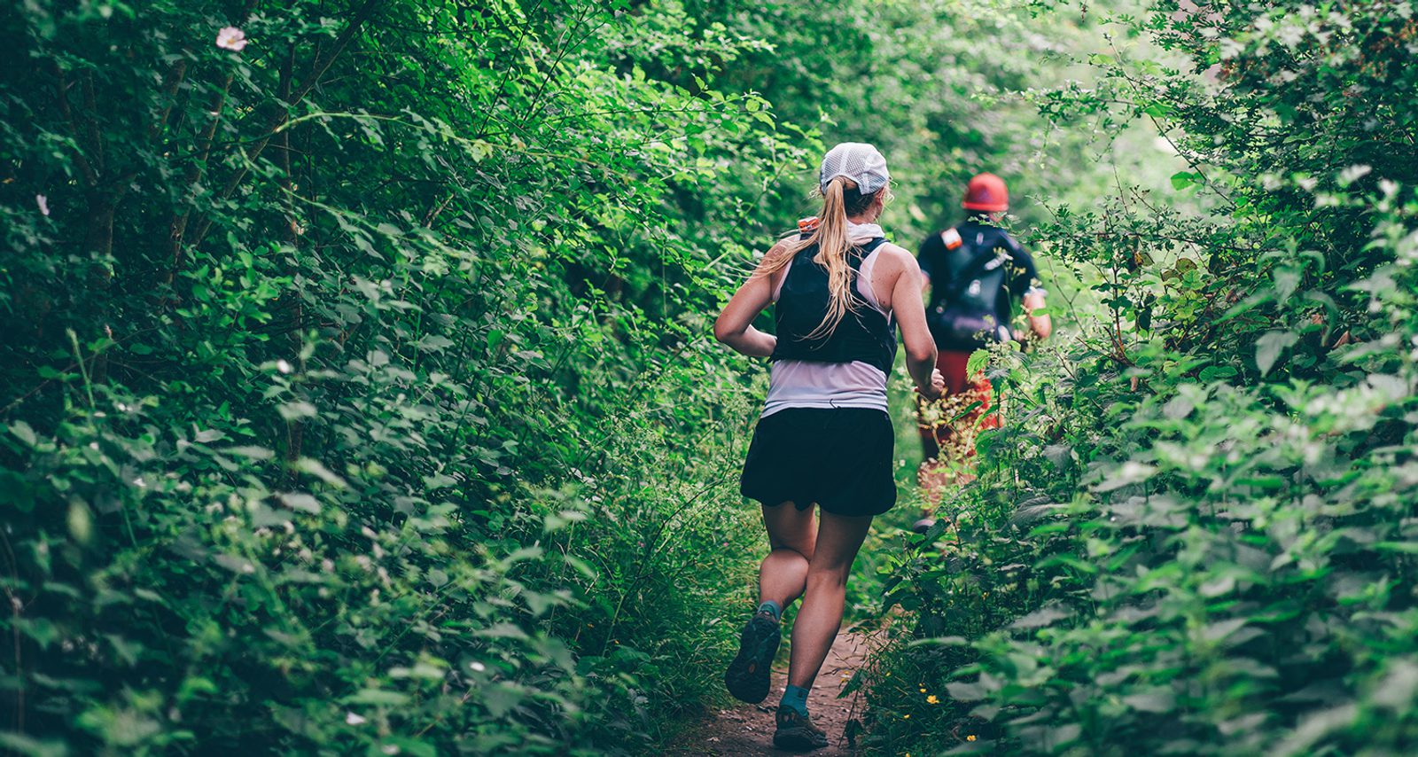 Two people run on a narrow dirt trail through lush green forest, surrounded by dense foliage. The runner in the foreground wears a white cap, black shorts, and a tank top. The second runner is farther ahead on the path.