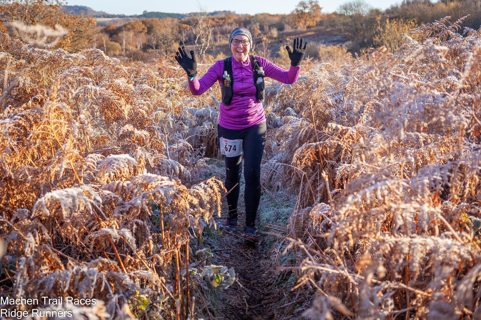 A person dressed in outdoor running gear, including a purple jacket and black pants, jogs along a narrow frosty trail surrounded by brown ferns. They are smiling, wearing gloves and a hat, and have their hands raised. Text reads "Machen Trail Races, Ridge Runners" in the bottom left corner.