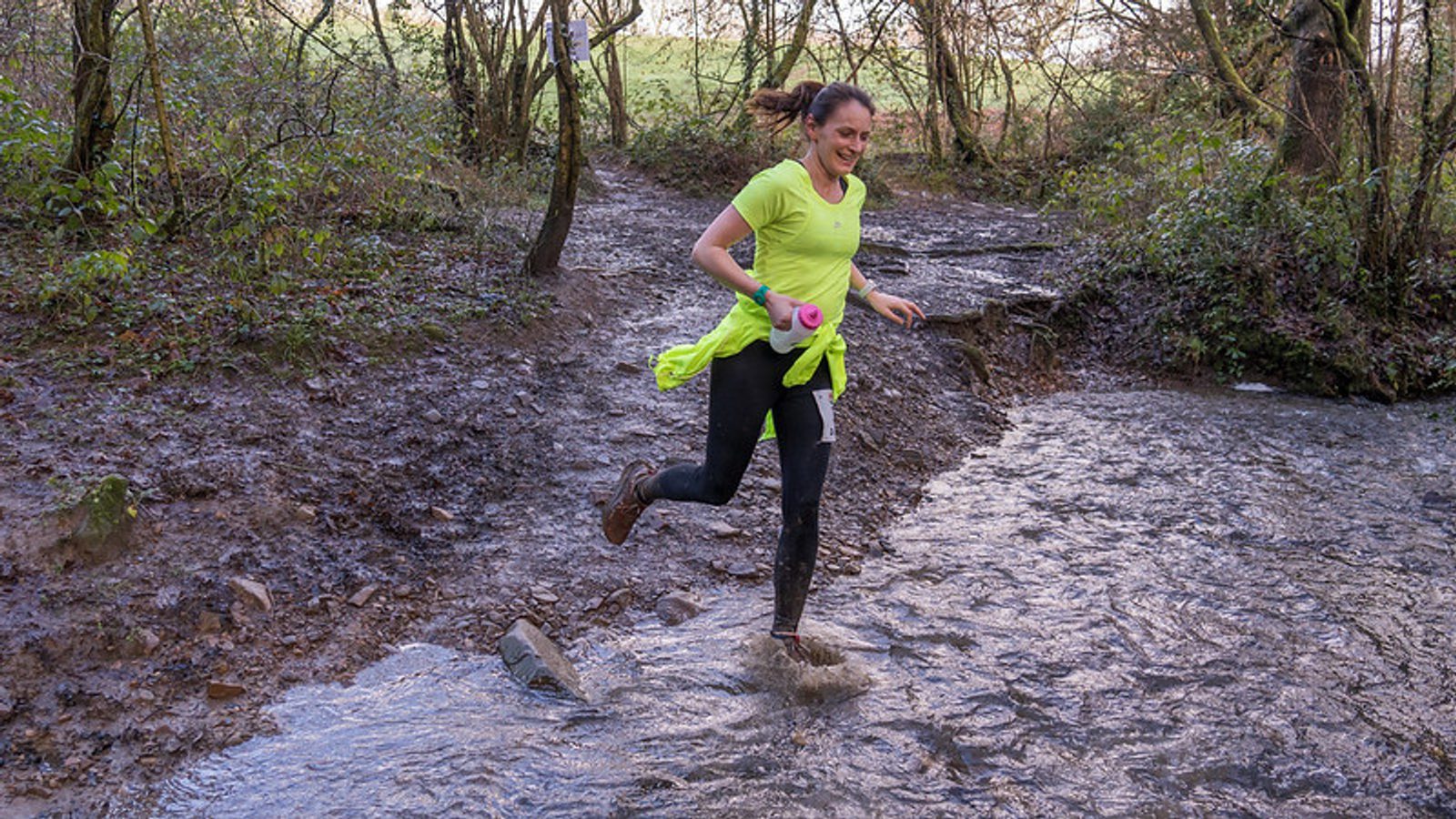 A woman wearing a bright yellow shirt and black leggings is running through a muddy trail in a forest. She is smiling and appears to be in the middle of a crossing a shallow stream, splashing water around her. She holds a pink water bottle in one hand.