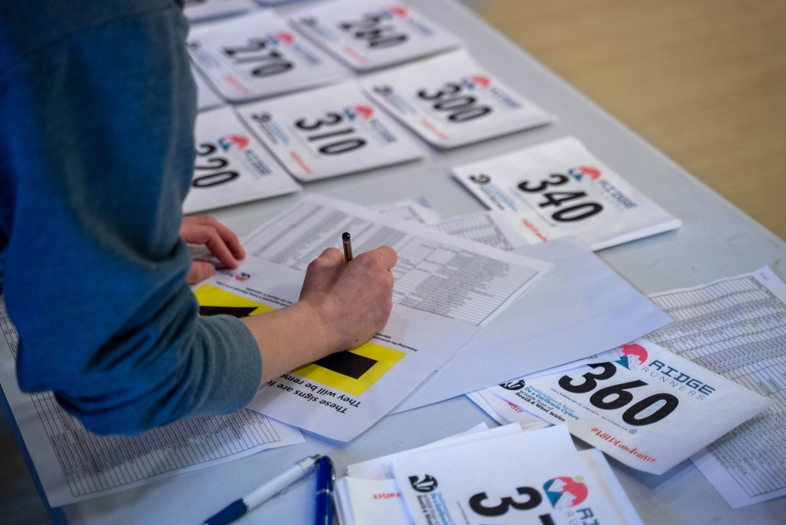A person is filling out a form on a table covered with race bibs displaying numbers such as 300, 340, and 360. There are also multiple pens and a list with printed text visible on the table. The scene suggests preparation for a race or sporting event.