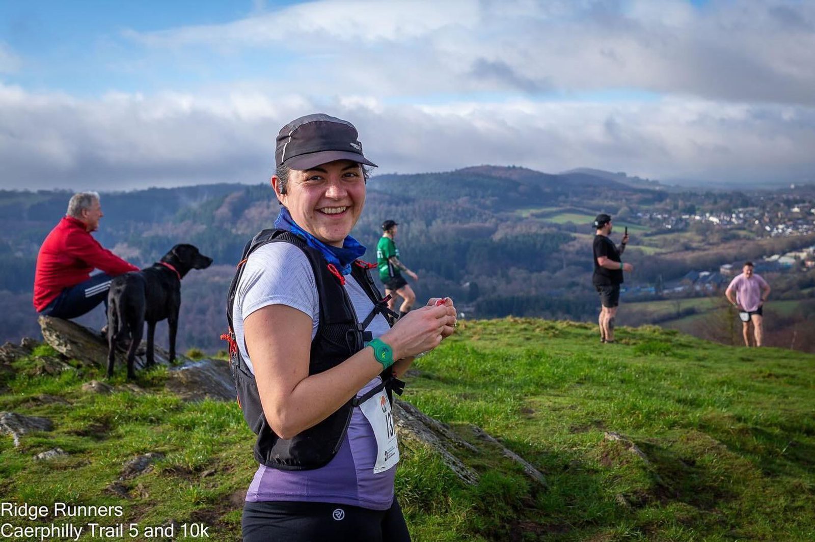 A woman wearing a cap and running gear smiles at the camera atop a grassy hill with a scenic valley and mountains in the background. Several other people and a black dog are nearby, enjoying the view. Visible text: "Ridge Runners Caerphilly Trail 5 and 10k".