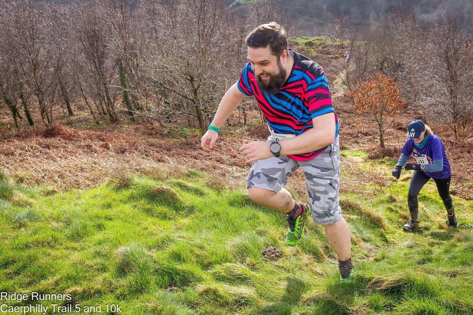 Two runners are pictured on a grassy hill amidst a natural, wooded backdrop. The foreground runner wears a striped shirt and is enthusiastically moving uphill. Behind, another runner in a purple top and number bib follows. The mood is energetic and focused.