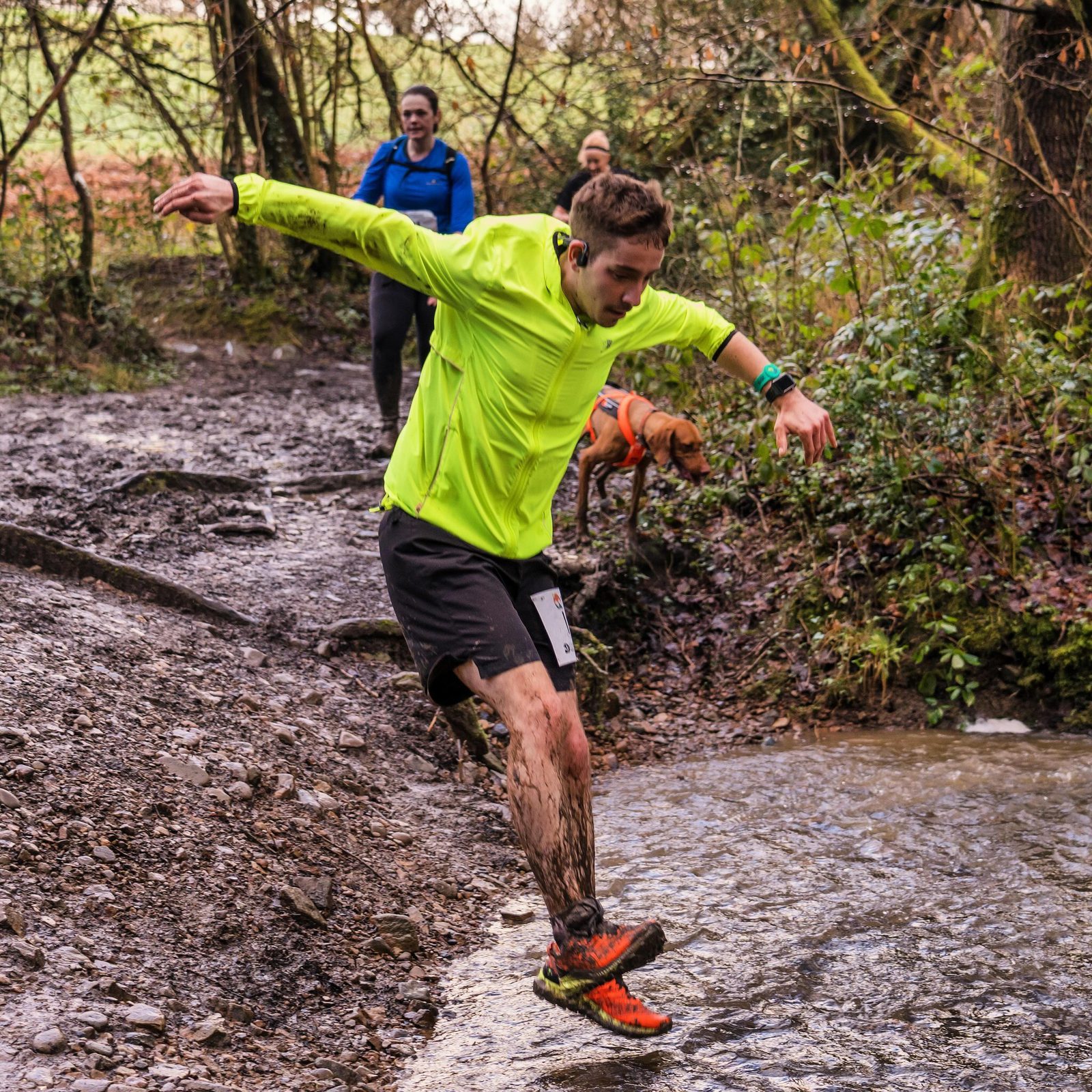 A man in a bright yellow jacket and black shorts is leaping over a small stream on a muddy trail. His shoes and legs are muddy, and he seems to be focusing on his jump. In the background, another runner and a dog approach the stream.