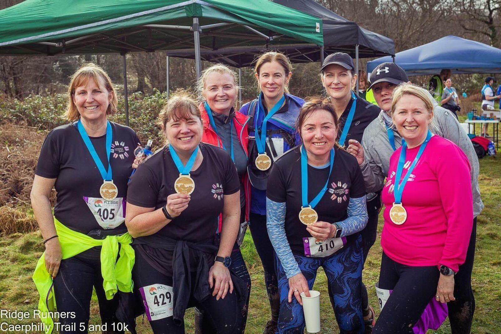A group of eight runners, all women, pose proudly with their medals after completing a trail race. They are dressed in athletic wear and are smiling at the camera. Tents and other participants are visible in the background. Text reads "Ridge Runners Caerphilly Trail 5 and 10k.