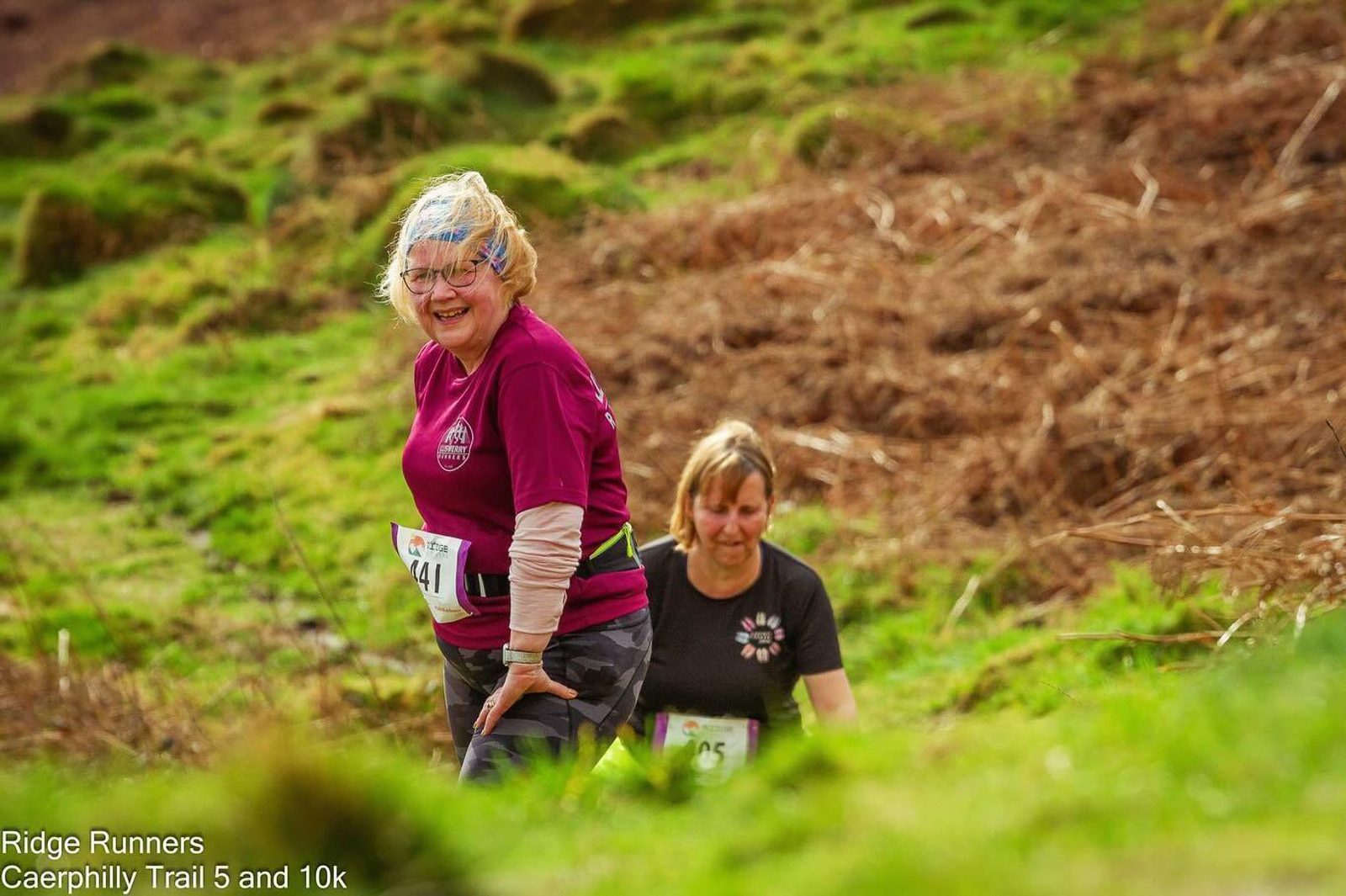 Two women are participating in an outdoor trail run through a grassy and hilly landscape. Both are wearing race bibs and athletic clothing. The woman in front is smiling and appears to be in high spirits. Text in the image reads "Ridge Runners, Caerphilly Trail 5 and 10k.