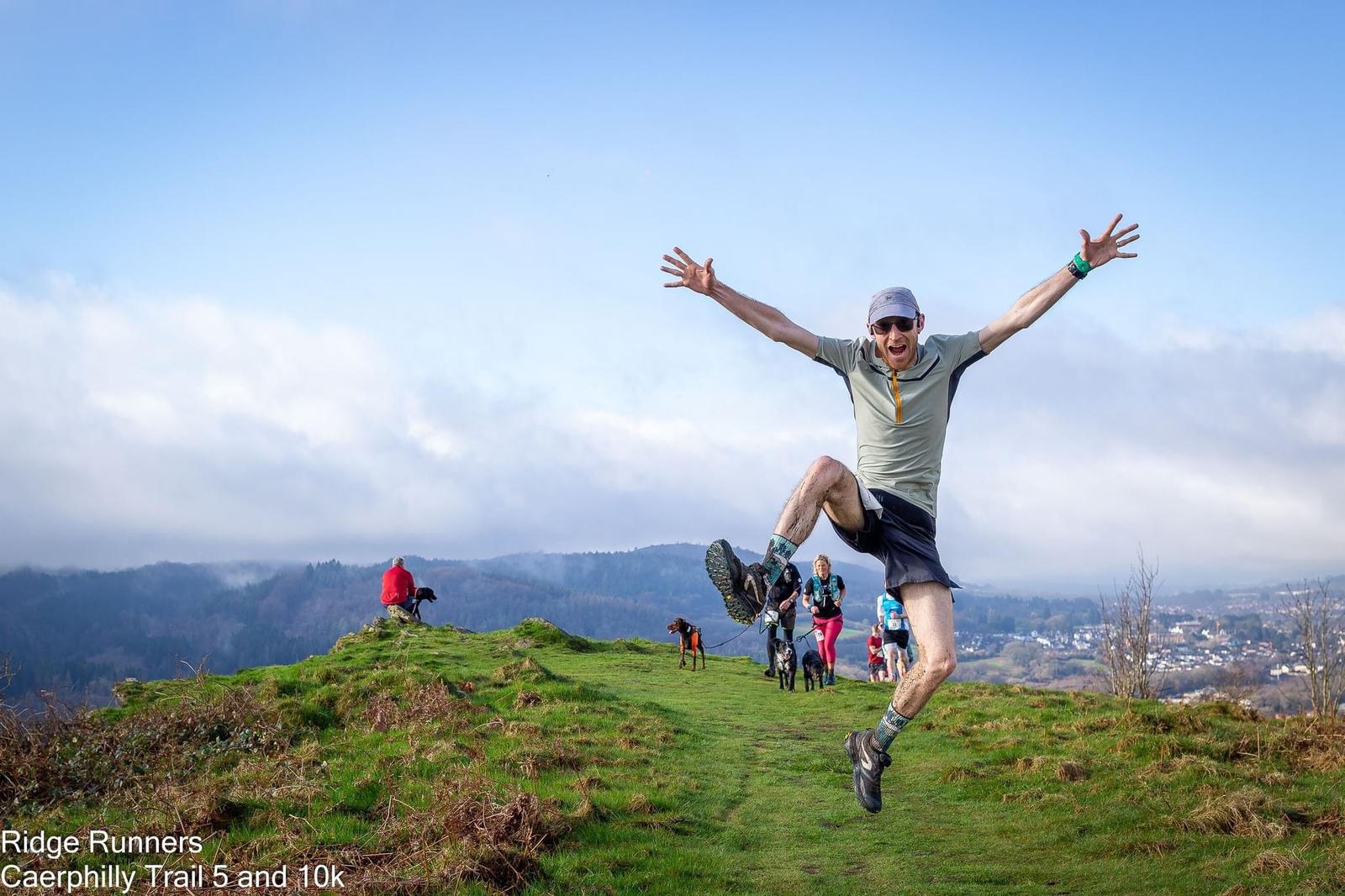 A man is mid-jump with arms and legs spread wide on a grassy trail on a hilltop. He wears a cap, sunglasses, a gray shirt, and shorts. Other people and dogs are on the trail in the background. Text on the image reads: Ridge Runners Caerphilly Trail 5 and 10k.