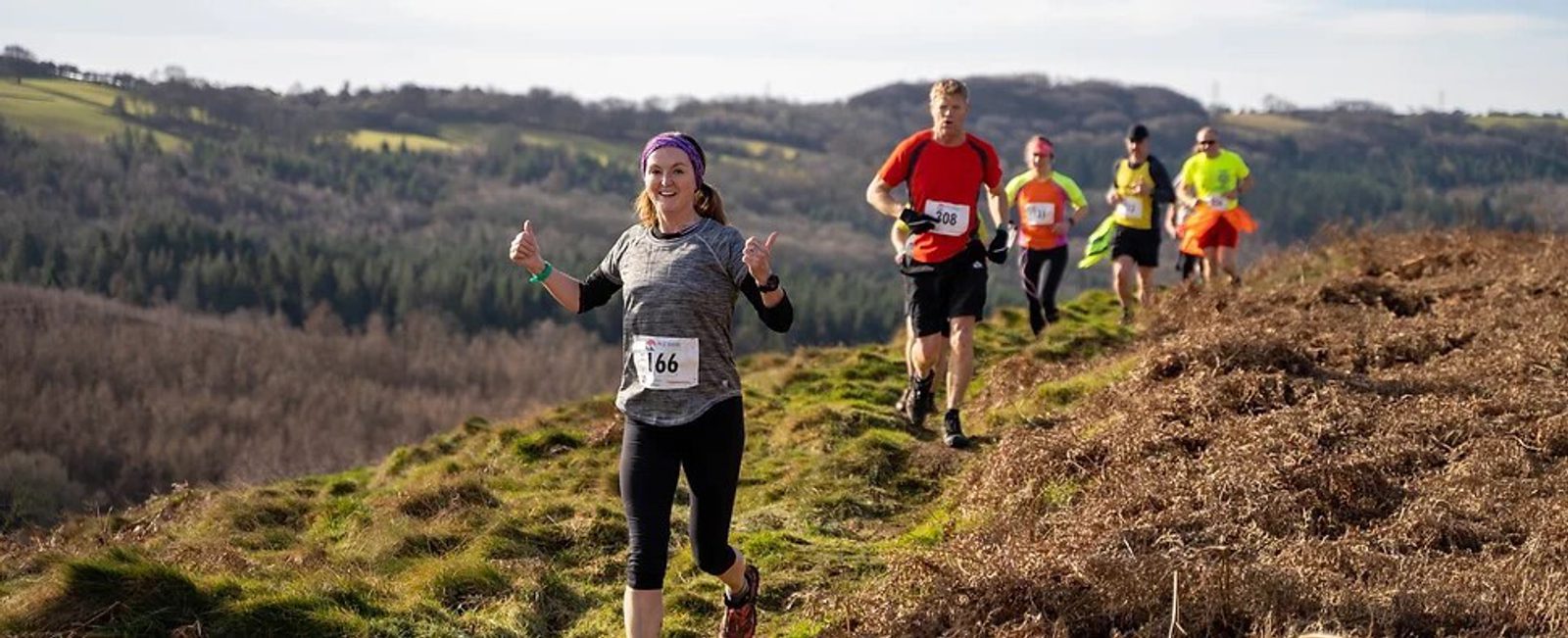 A group of runners participates in a trail race on a narrow path through a scenic, hilly landscape with lush greenery. The runner closest to the camera, wearing a grey shirt and a purple headband, gives a thumbs-up while others follow closely behind.