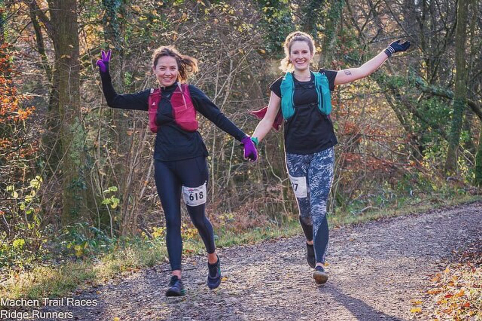 Two women in athletic gear run on a wooded trail, smiling and holding hands. Both wear numbered race bibs, one with the number 618 and the other with 77. They have hydration backpacks and are surrounded by autumn foliage. The woman on the left flashes a peace sign.