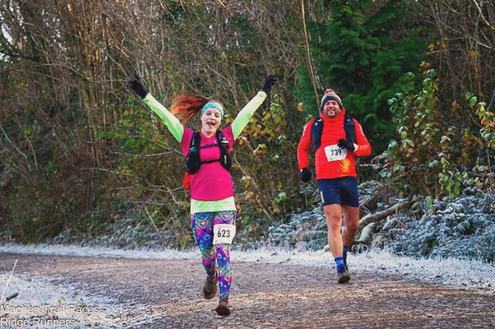 Two runners on a forest trail celebrate mid-race. The foreground runner, wearing colorful leggings and raising their arms in excitement, is slightly ahead of another runner in a red jacket and black shorts. Both wear numbered bibs and backpacks.