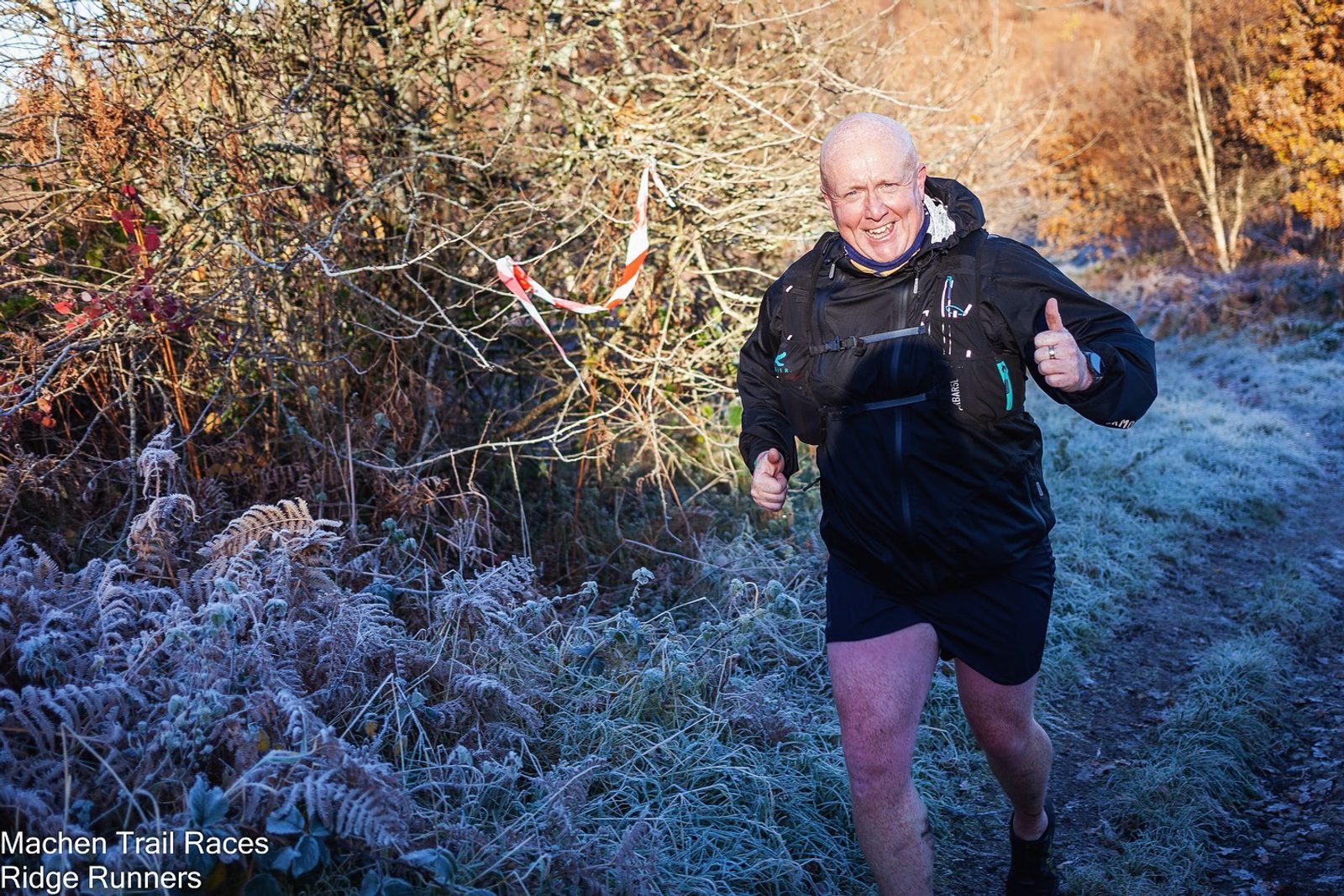 A man in outdoor running gear smiles and gives a thumbs-up while running along a frosty, narrow trail surrounded by autumnal trees and bushes. He is participating in Machen Trail Races, as indicated by the text "Machen Trail Races" and "Ridge Runners" on the bottom-left corner.