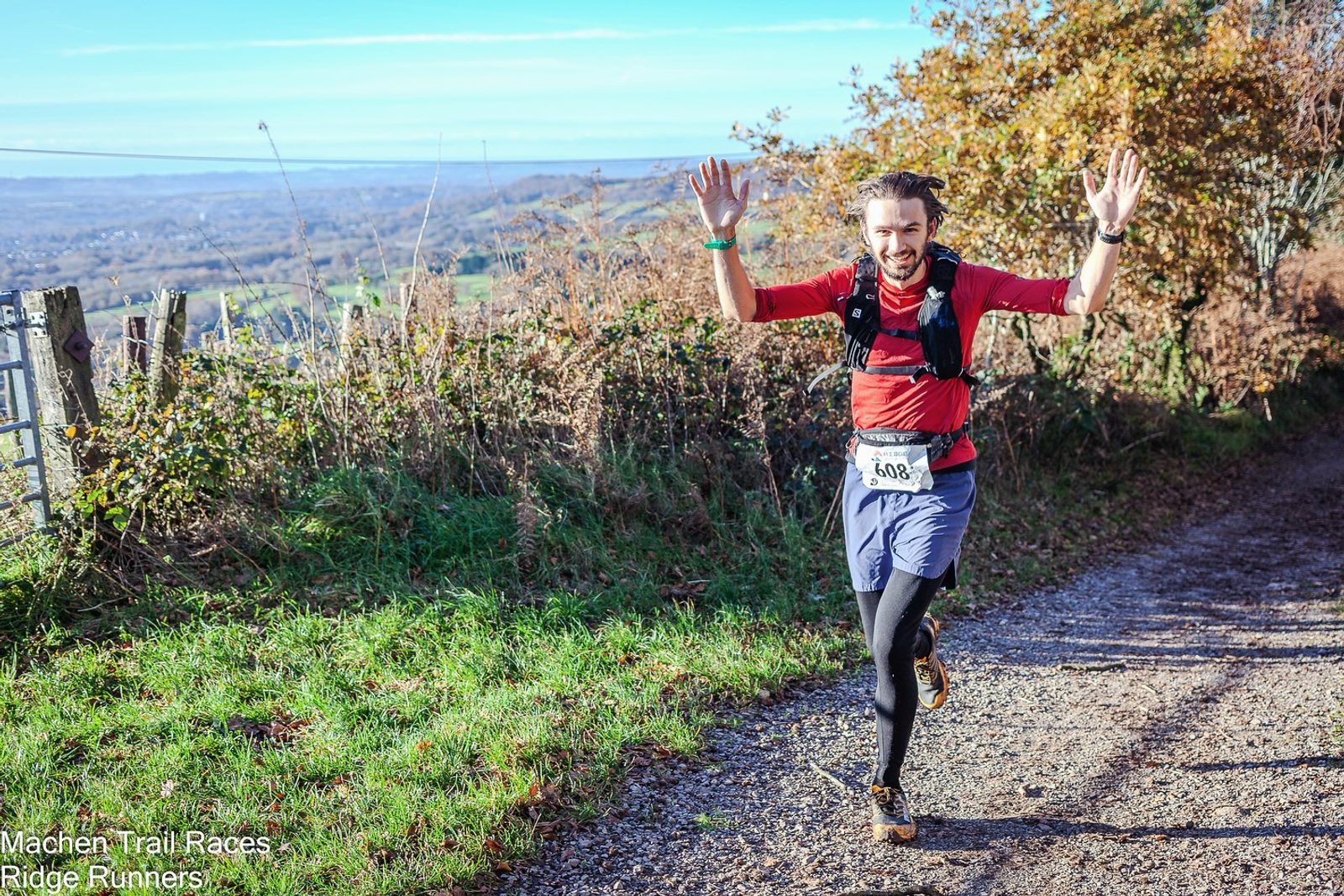 A runner in a red shirt and black leggings, wearing a race number "488", smiles and raises both arms while running on a scenic trail. The path is surrounded by greenery and autumn trees. The background reveals a wide landscape under a clear blue sky.