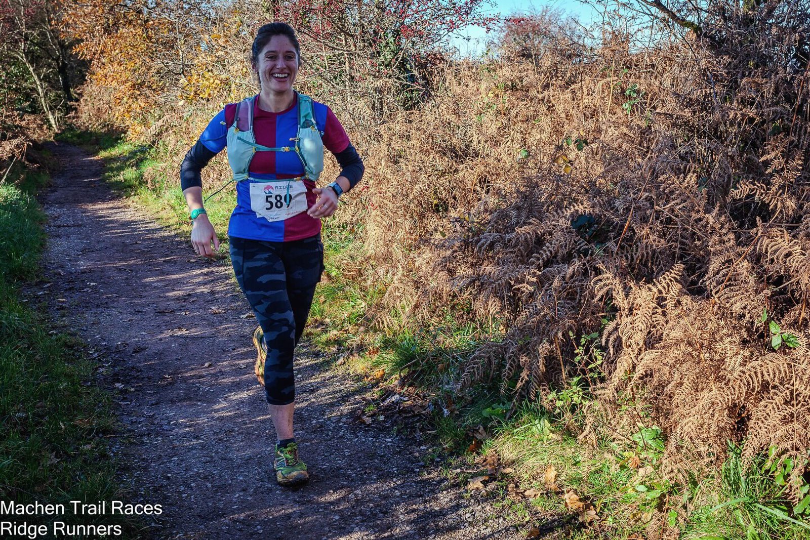 A person wearing a red top, black and grey camouflage leggings, a blue hydration pack, and race bib number 581 is running on a trail surrounded by autumn foliage. They are smiling and appear to be enjoying the race. Text in the bottom left corner reads "Machen Trail Races" and "Ridge Runners.
