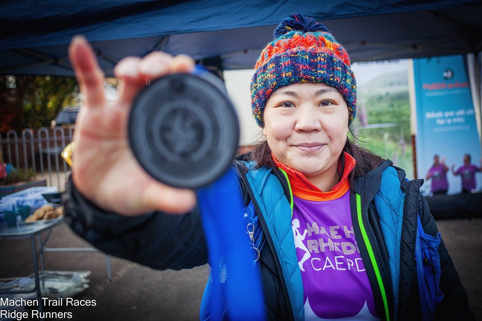 A person smiling and holding up a medal towards the camera. They are wearing a colorful knit hat, a blue and orange jacket, and a purple shirt with a logo. There is a tent and banners in the background. Text in the corner reads "Machen Trail Races Ridge Runners.