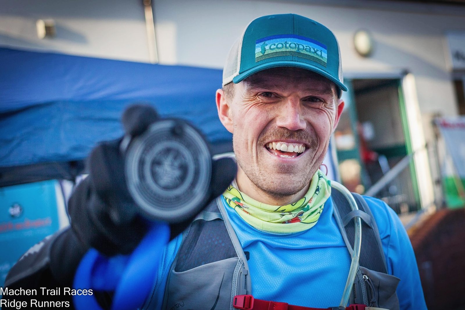 A smiling man wearing a blue shirt, gloves, and a baseball cap holds up a medal in front of him. He is outdoors, possibly at an event or race, as indicated by the background setup, and appears to be happy and celebrating an achievement. The text "Machen Trail Races Ridge Runners" is in the bottom left corner.