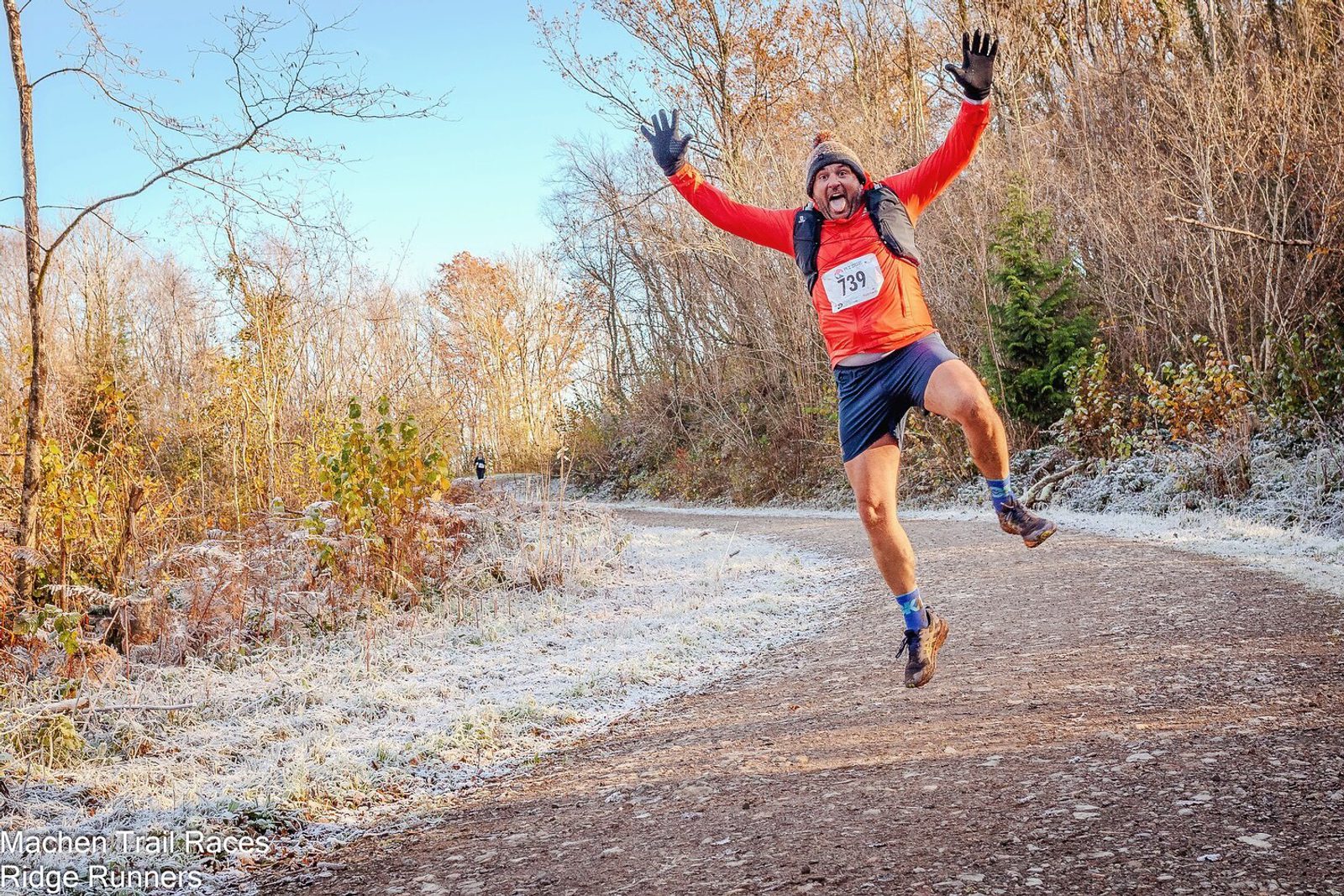 A joyful runner wearing an orange shirt, blue shorts, and a beanie jumps with arms raised while running on a frosty forest trail. The trail is surrounded by leafless trees and winter foliage. The runner has a race number 739 pinned to their shirt.