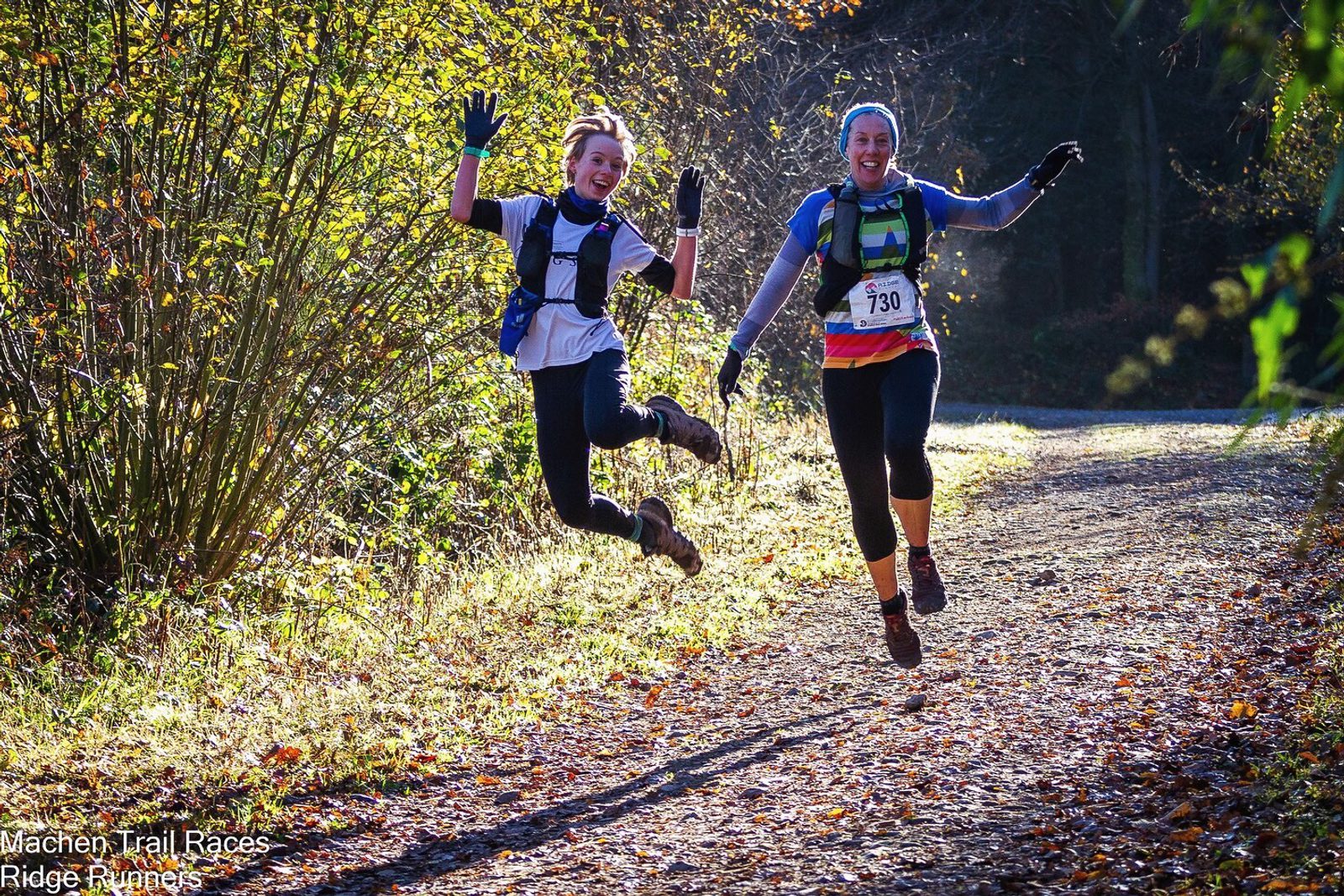 Two runners, wearing race bibs, happily jump in the air while running on a sunlit forest trail. The person on the left has their arms raised high, while the person on the right smiles widely. Autumn leaves blanket the ground, and the surrounding foliage glows in the sunlight.